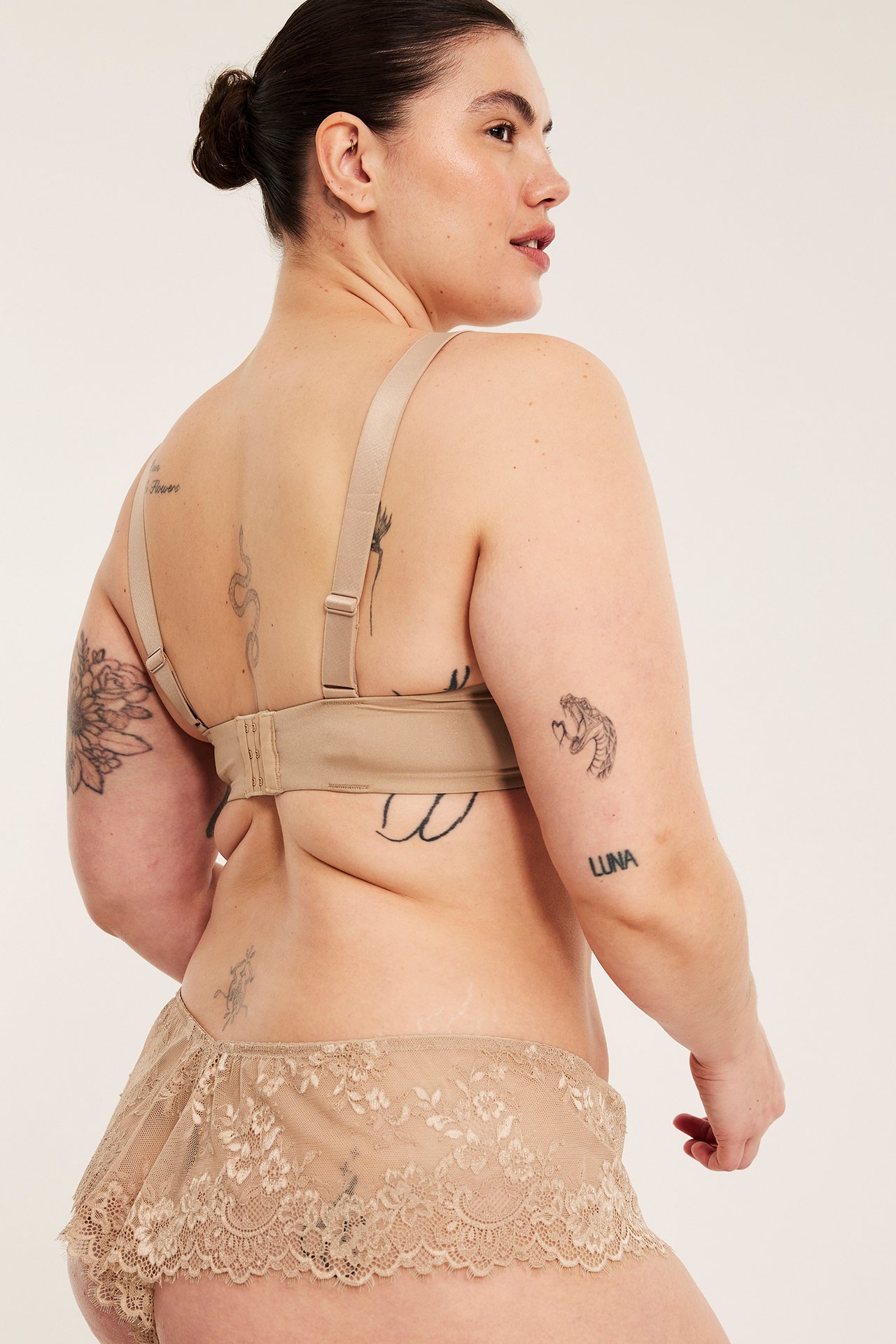 Nainen selin, yllään korkeavyötäröiset beiget pitsiset alushousut ja yhteensopivat beiget rintaliivit.