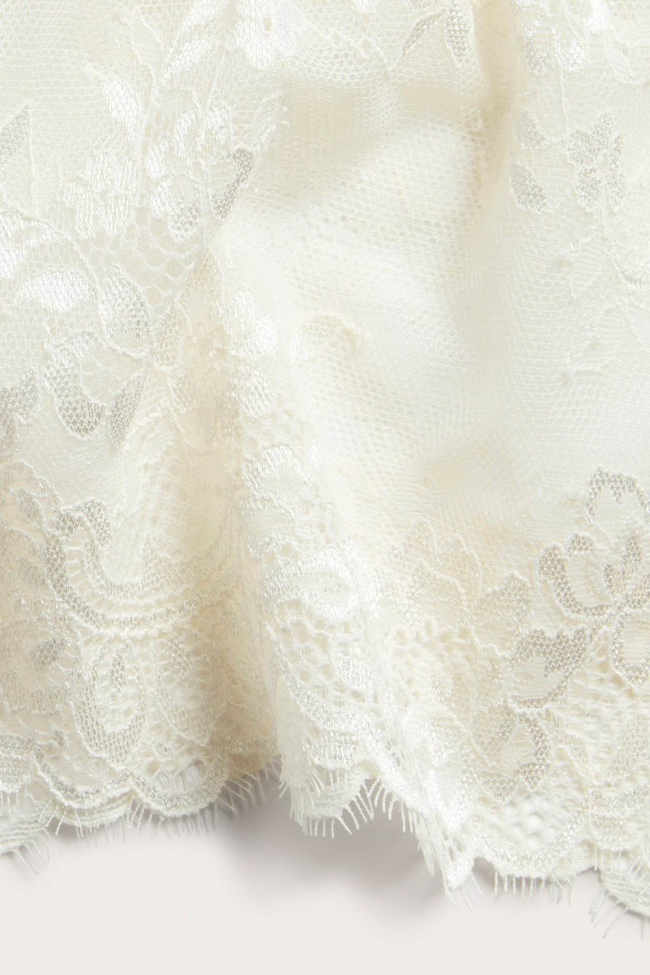 Zbliżenie na ecru koronkowy materiał z ozdobnym kwiatowym wzorem i falbankowym brzegiem.