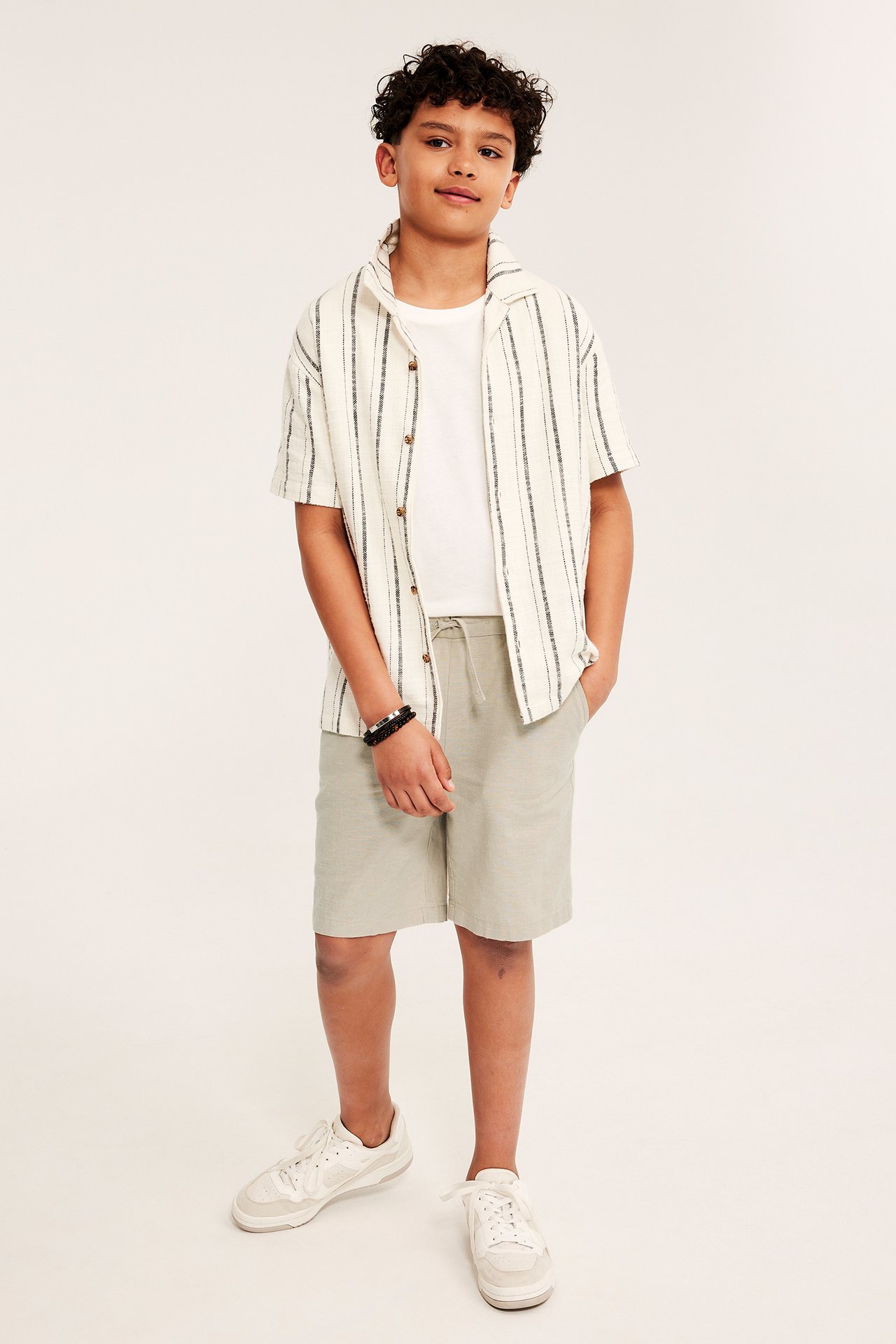 Framifrån: Barn bär beiga linneshorts, öppen randig skjorta över vit t-shirt, vita sneakers.