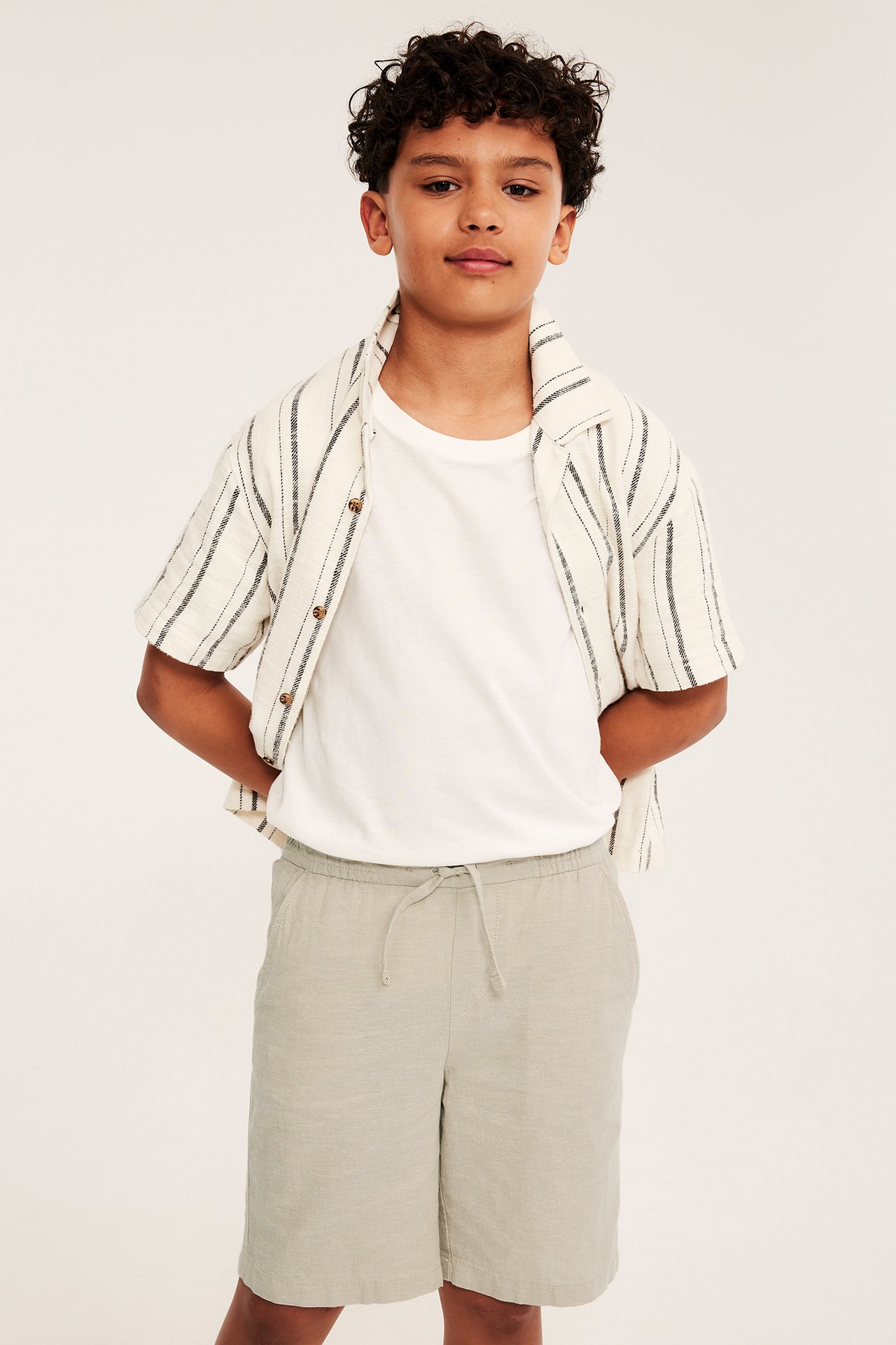 Framifrån: ett barn i beigefärgade linneshorts med dragsko, en vit t-shirt och en öppen randig skjorta.
