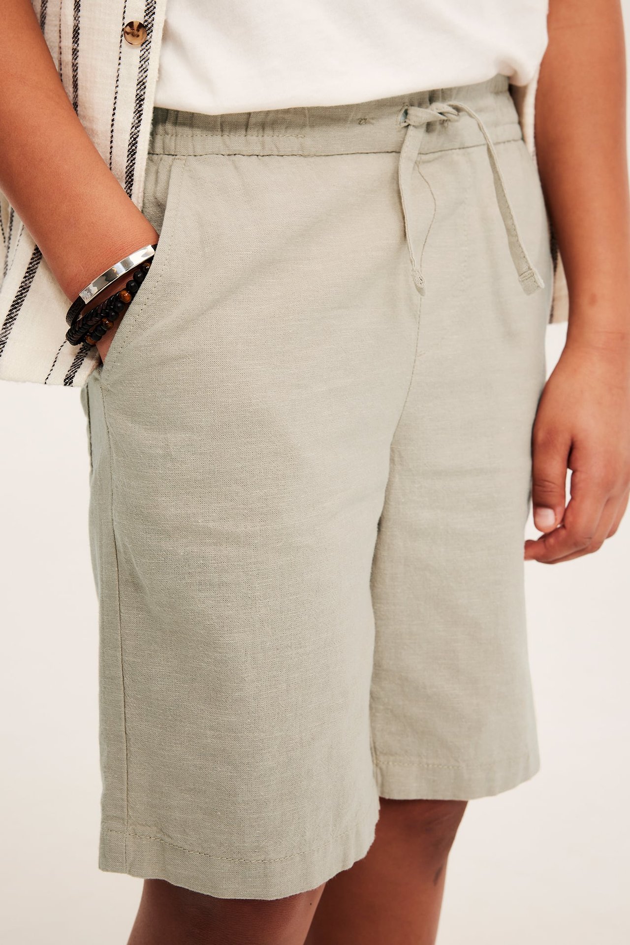 Närbild på ett barn i ljusbeige linneshorts med resår i midjan, en vit t-shirt och randig skjorta.