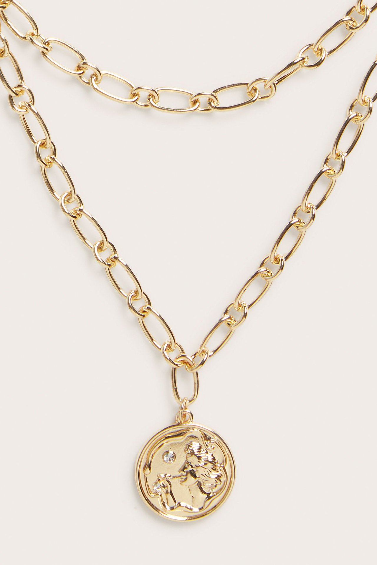 Lähikuva kultaisesta kaulakorusta, jossa kaksi ketjua ja pyöreä riipus kohokuvioinneilla sekä pienillä kivillä.