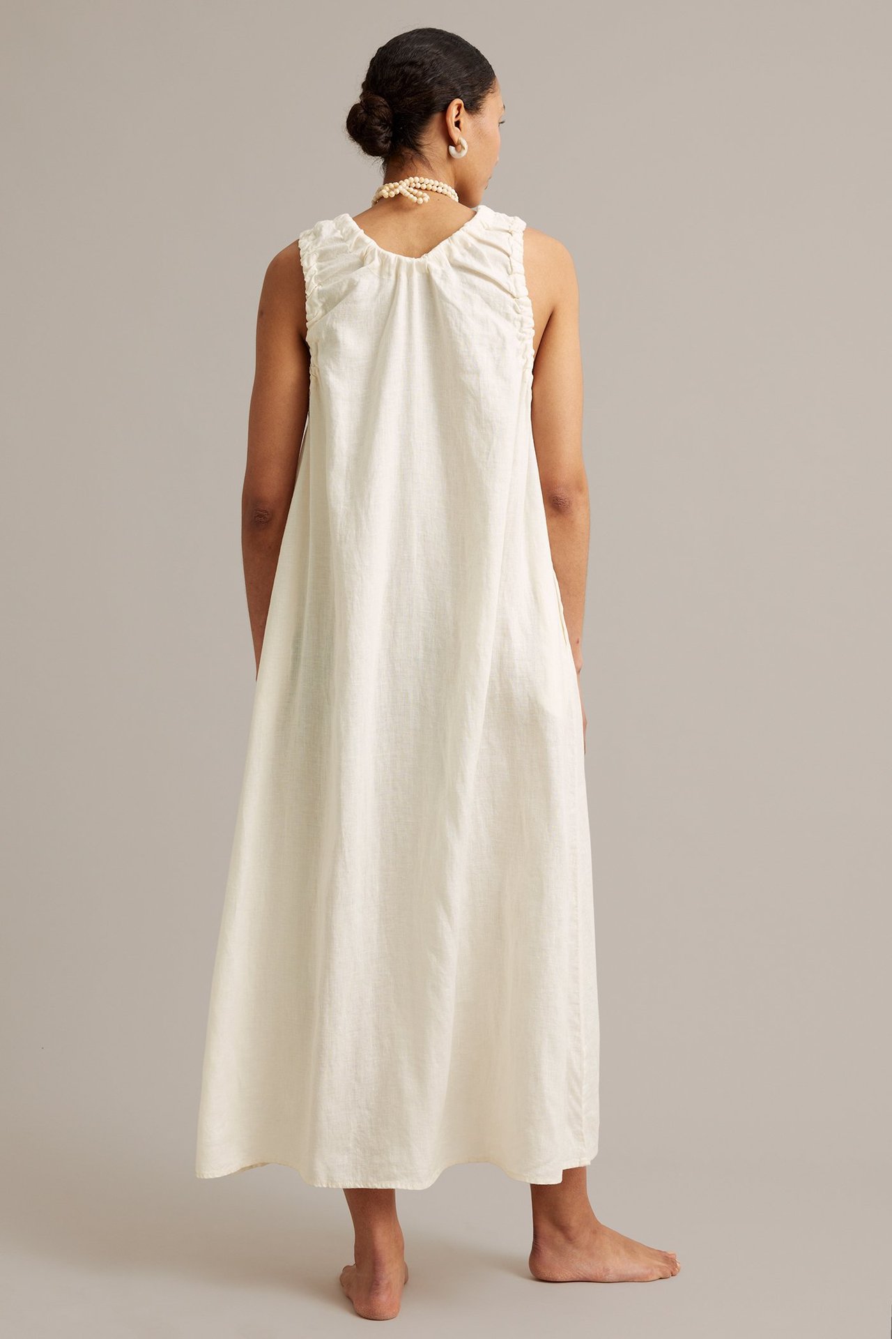 Widok z tyłu kobiety w białej lnianej sukience maxi z marszczonym dekoltem, z perłami i kolczykami.