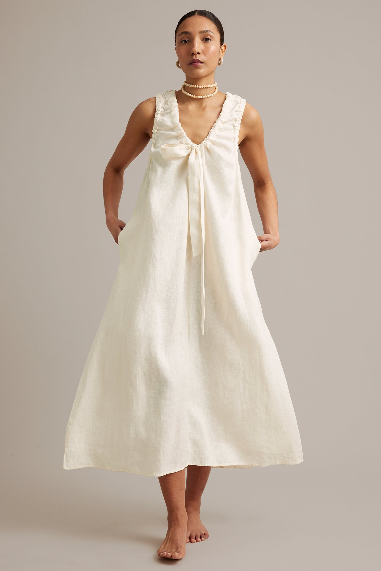 Framifrån: Kvinna i vit ärmlös linneklänning med knyt i V-ringning, sidfickor och pärlhalsband.