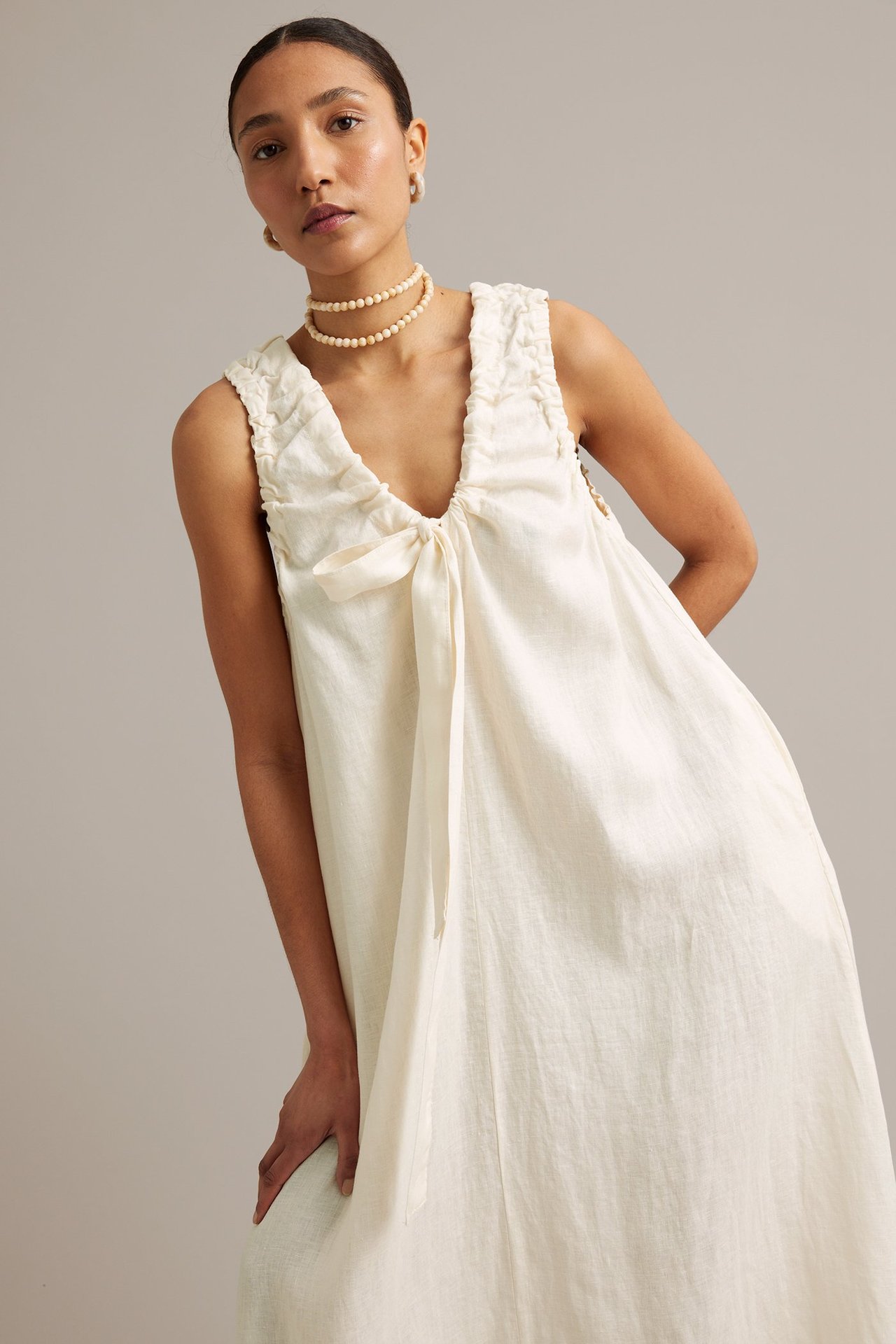 Kobieta w białej, lnianej sukience bez rękawów z dekoltem V, wiązaniem i naszyjnikiem z pereł.