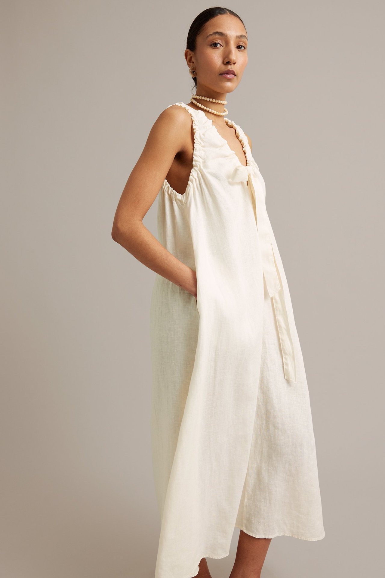 Z przodu: Kobieta w białej lnianej sukience bez rękawów z dekoltem V i wiązaniem. Nosi perłowy naszyjnik.