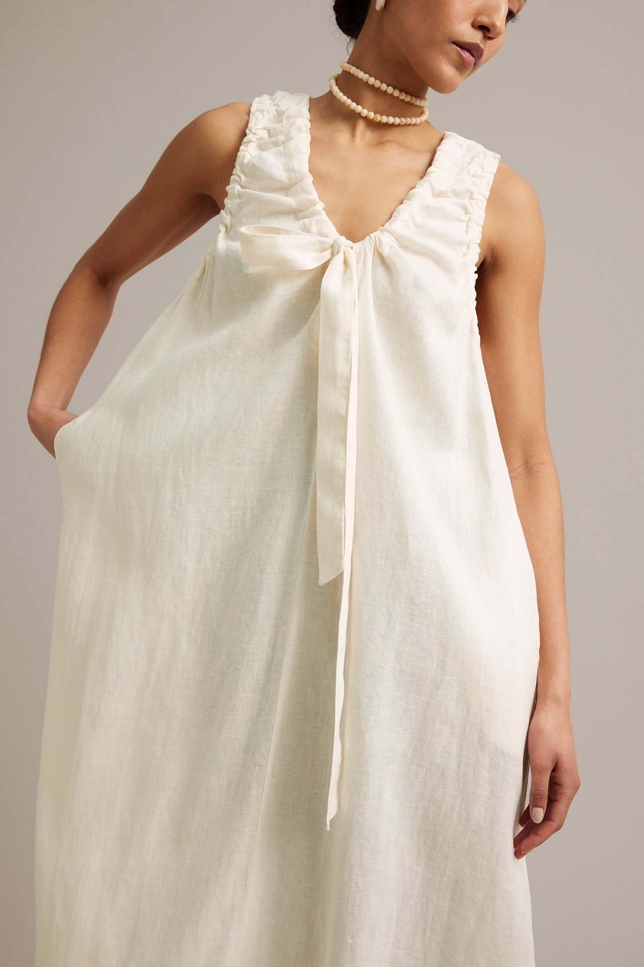Framifrån: Kvinna i vit ärmlös linneklänning med rynkad V-ringning, knytband, sidfickor och pärlhalsband.