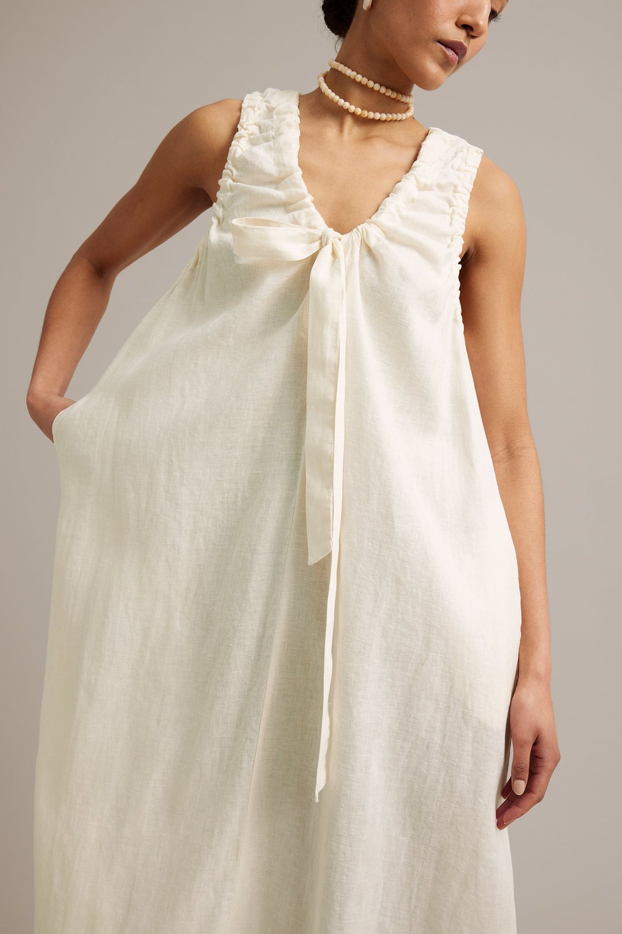 Edestä: Nainen valkoisessa hihattomassa pellavamekossa, jossa rypytetty V-pääntie, solmittava yksityiskohta, sivutaskut ja helmikaulakoru.