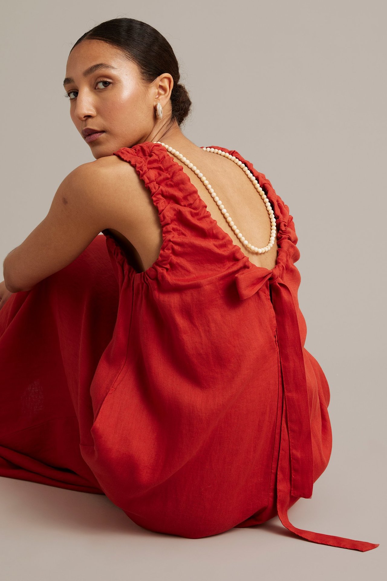 Kvinnas ryggvy i röd lös linneklänning med rynkade axlar, knyt baktill och ett pärlhalsband.