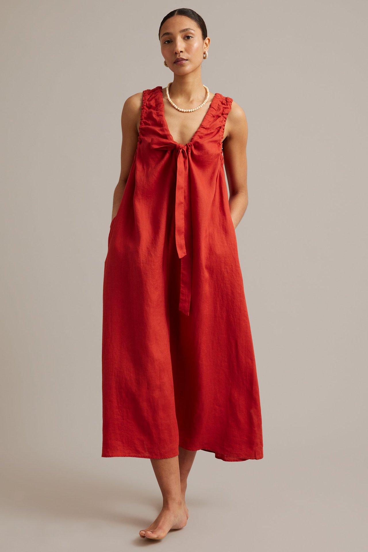 Widok z przodu: Kobieta w czerwonej lnianej sukience bez rękawów z dekoltem w serek, wiązaniem i marszczeniami. Nosi perły.
