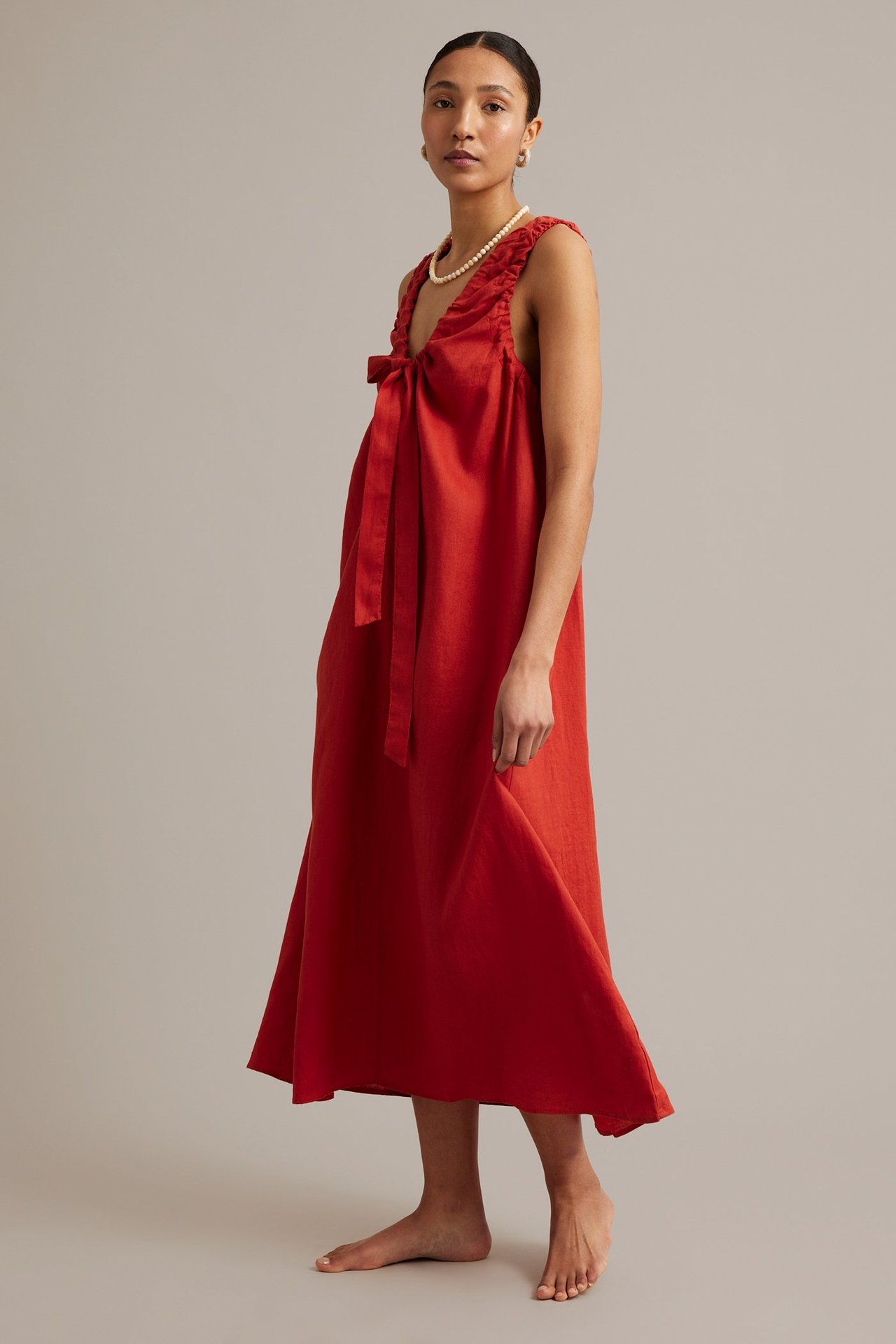 Framifrån, kvinna i lös röd linneklänning med knytband i V-ringningen, pärlhalsband och örhängen.