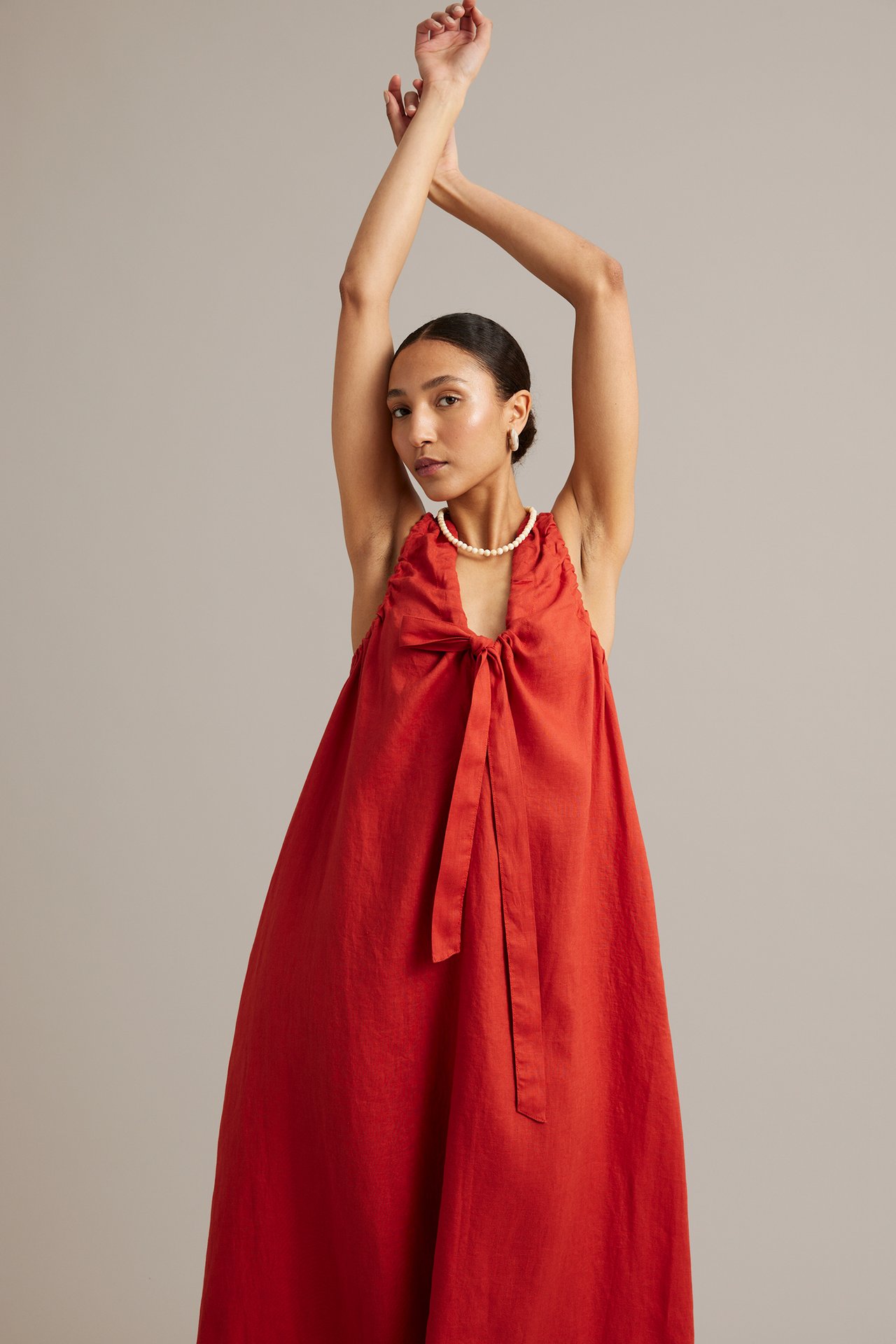 Framifrån: kvinna i en röd ärmlös linneklänning med knytning fram, volanger, pärlhalsband och ringörhängen.