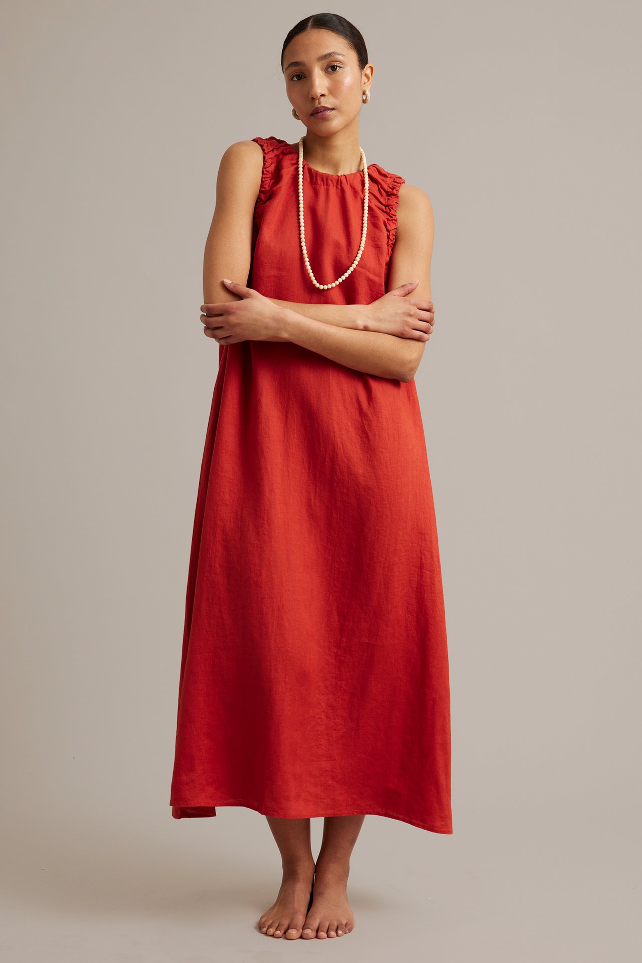 Framifrån: kvinna i röd ärmlös linneklänning med rynk, pärlhalsband och guldörhängen.