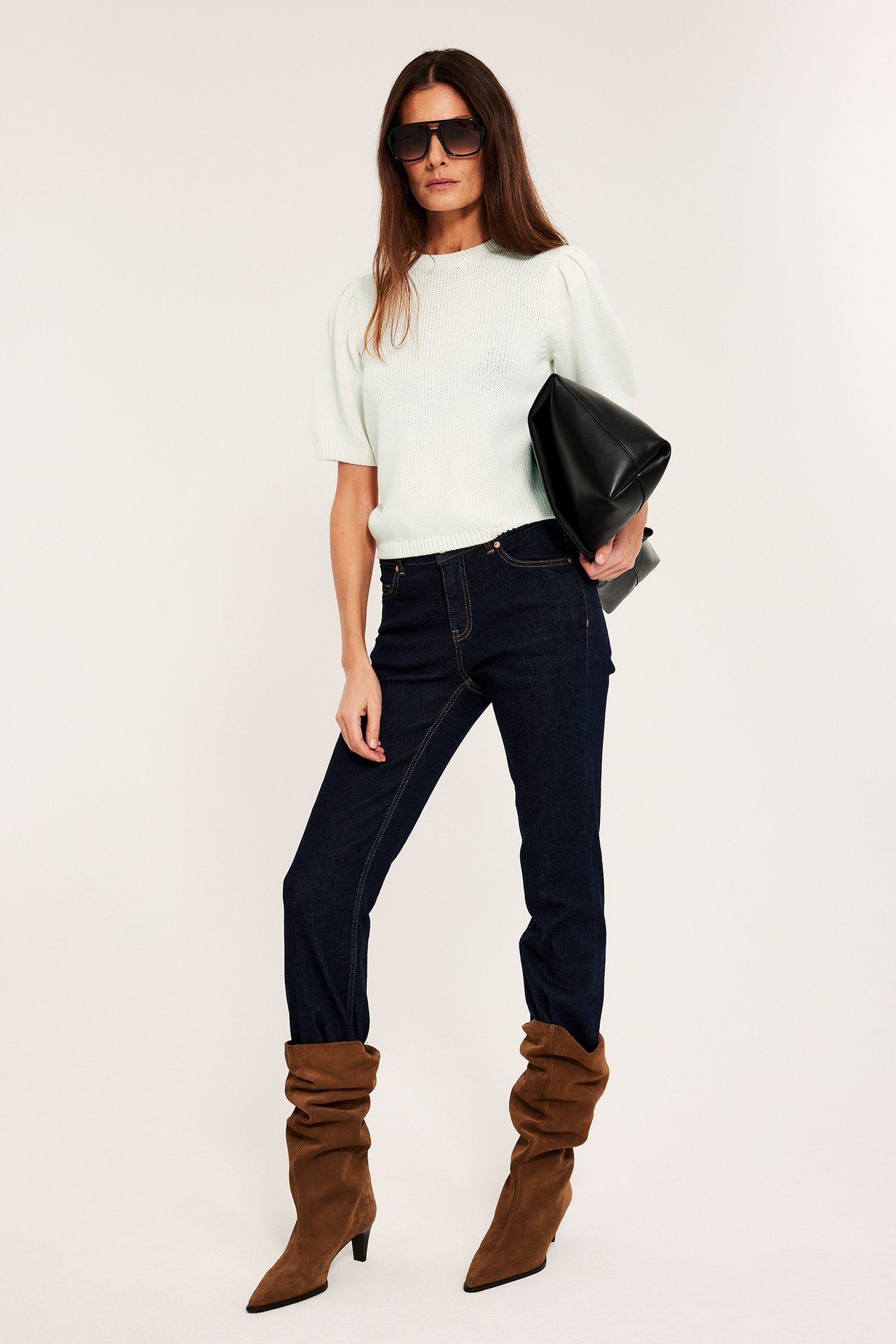 Framifrån: Kvinna i offwhite stickad bomullströja med puffärm, mörka jeans, bruna stövlar och svart clutch.