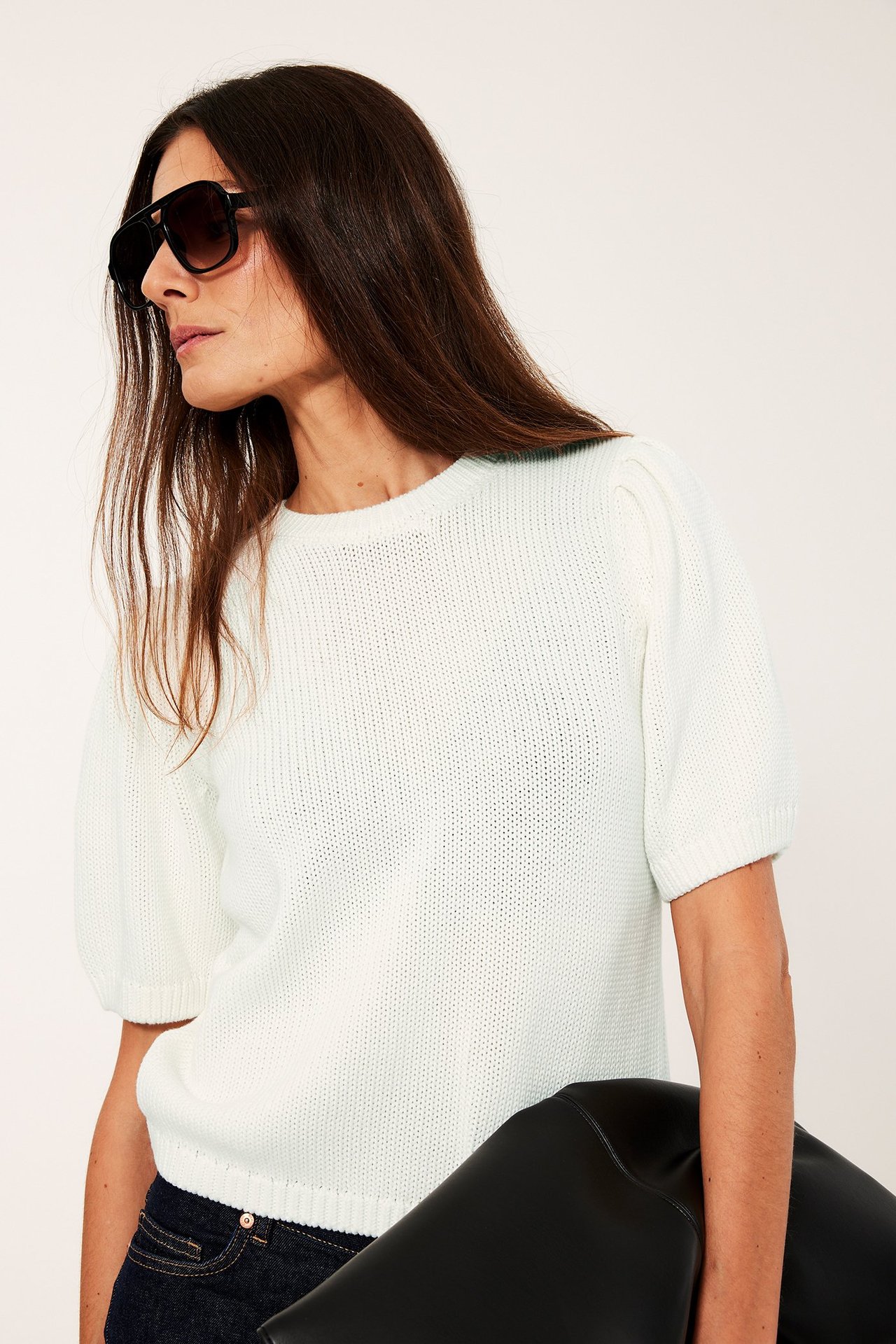 Sidovy av kvinna i off-white bomullsstickad tröja med puffärm, svarta solglasögon, håller svart clutch.