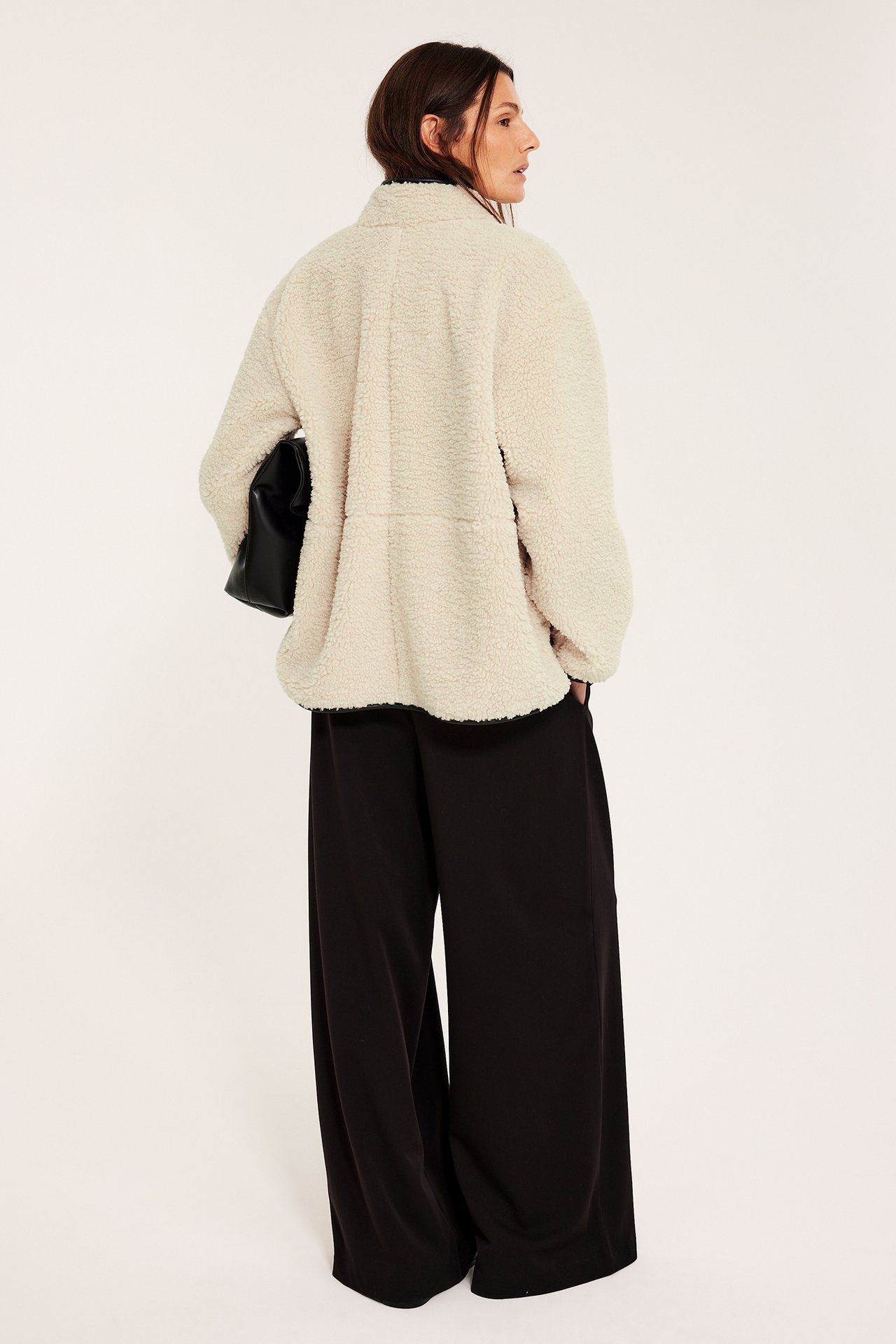 Ryggvy av kvinna i offwhite, avslappnad pilejacka, svarta vida byxor och en svart handväska.