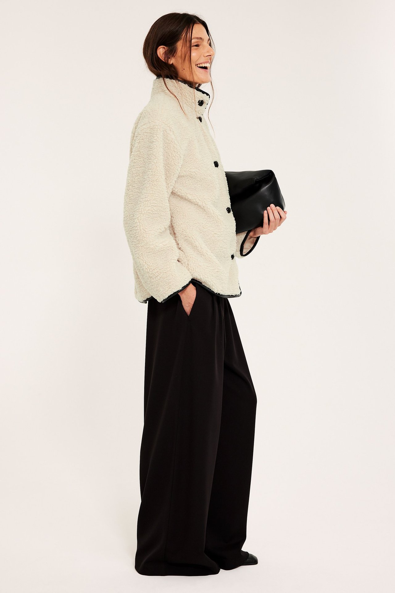 Sidovy: kvinna i offwhite pilejacka med svarta knappar, svarta vida byxor, håller i en svart clutchväska.
