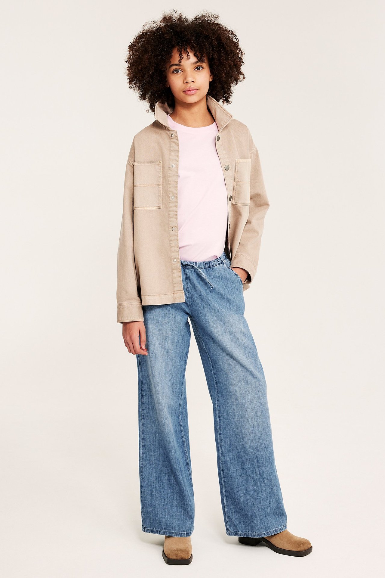 Forfra: barn i lyseblå myke denimbukser med snøring, lys rosa t-skjorte og beige overskjorte.