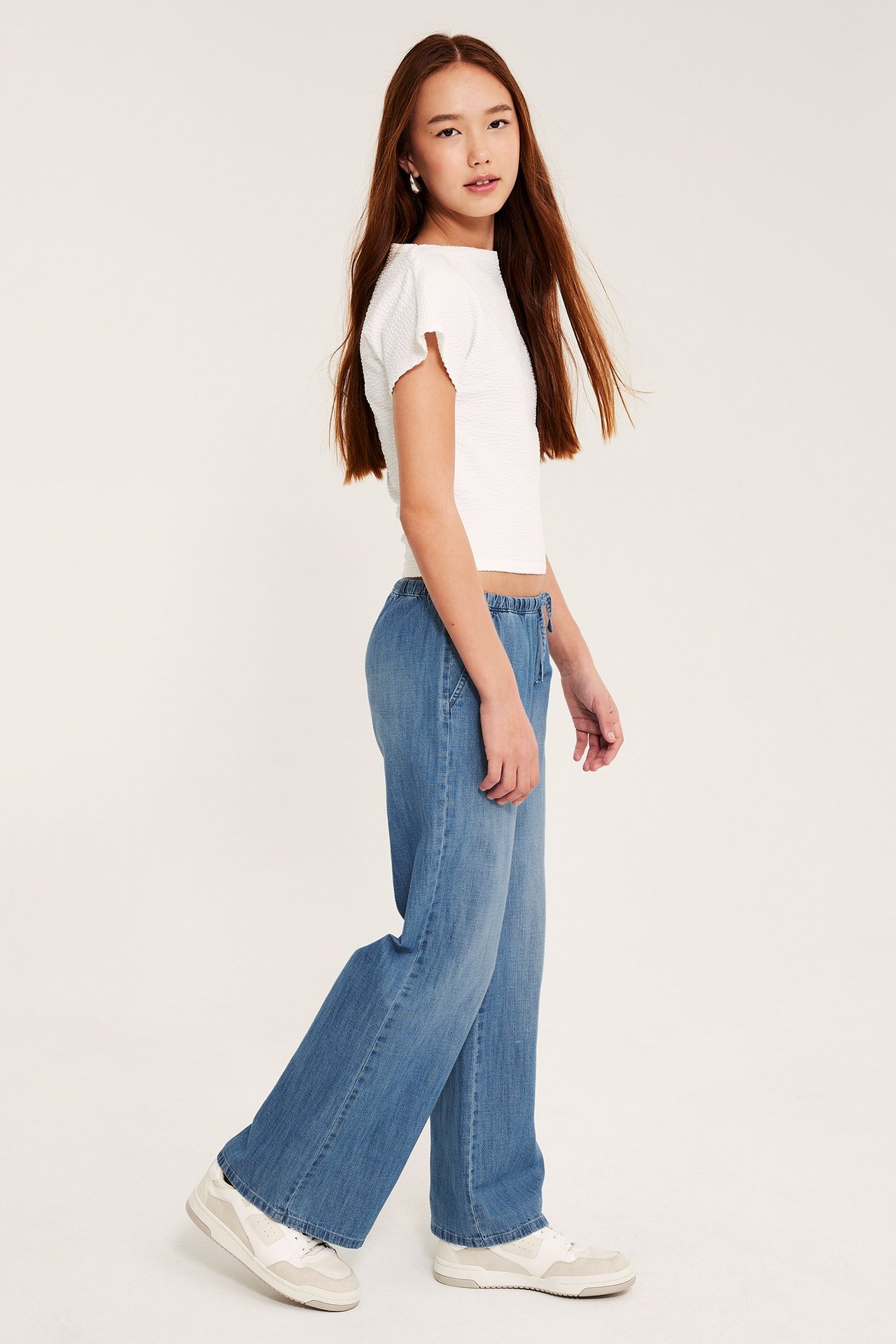 Sivukuva lapsesta vaaleansinisissä pehmeissä denimhousuissa kiristysnyörillä, valkoinen toppi ja tennarit.
