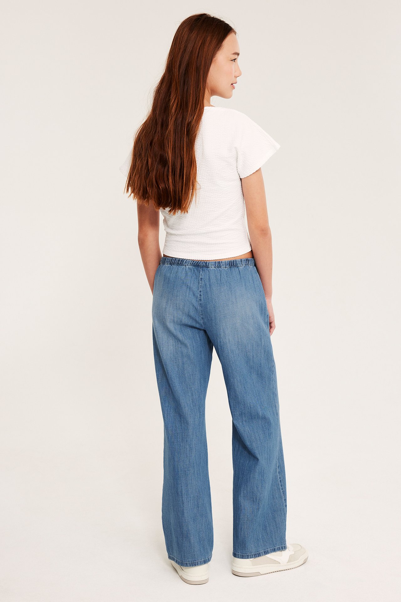 Ryggvy av ett barn i ljus denimbyxor med resår i midjan, en vit texturerad topp, och vita sneakers.