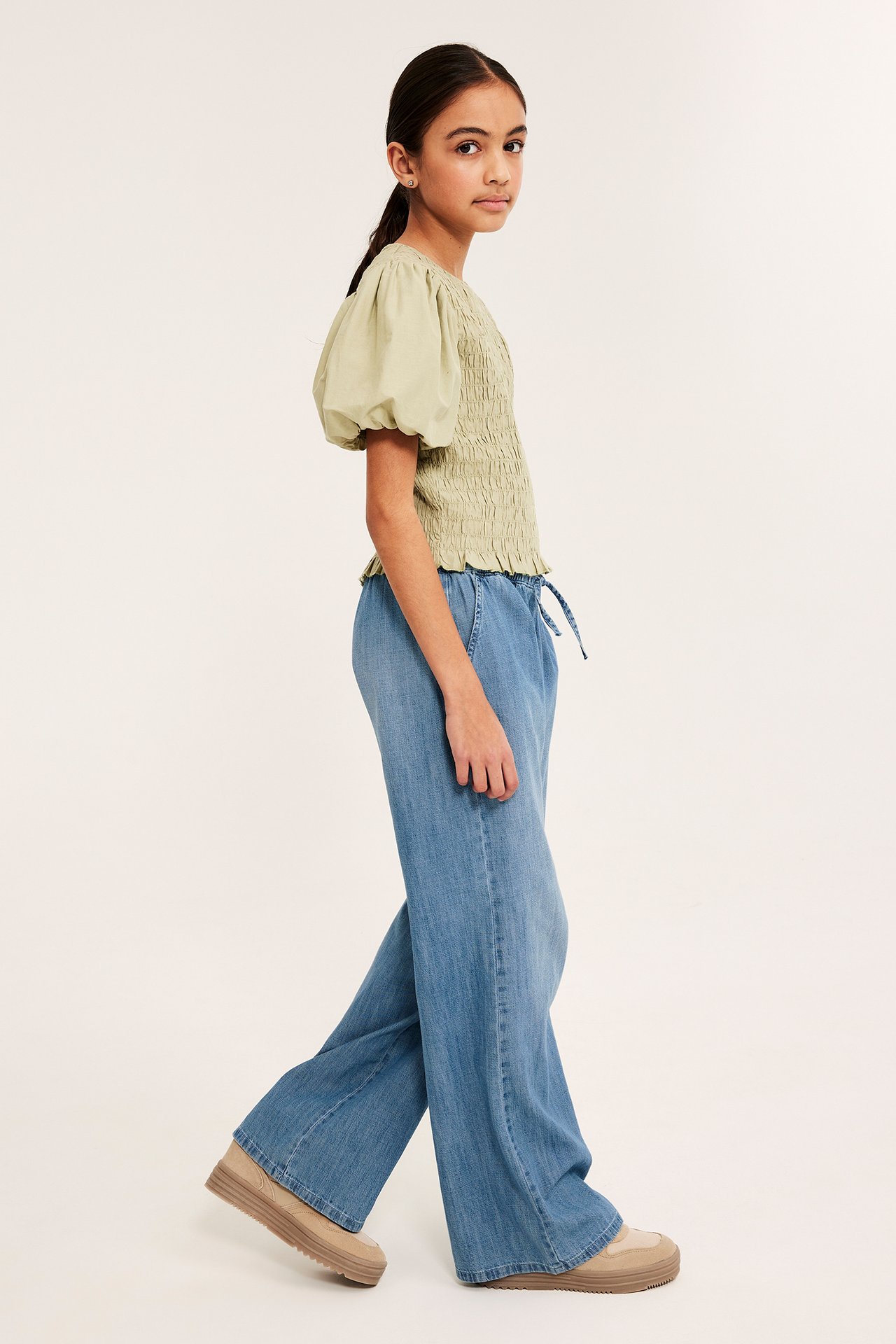 Sivukuva: lapsi yllään vaaleat farkkuhousut kiristysnyörillä, vihreä smokkipusero ja beiget lenkkarit.