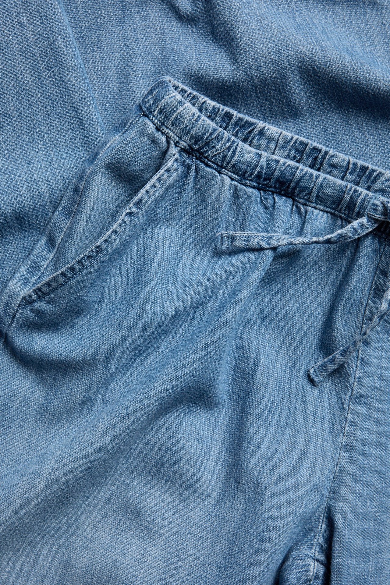 Nærbilde av lyseblå myke denimbukser med elastisk snøring i midjen og sidelommer.