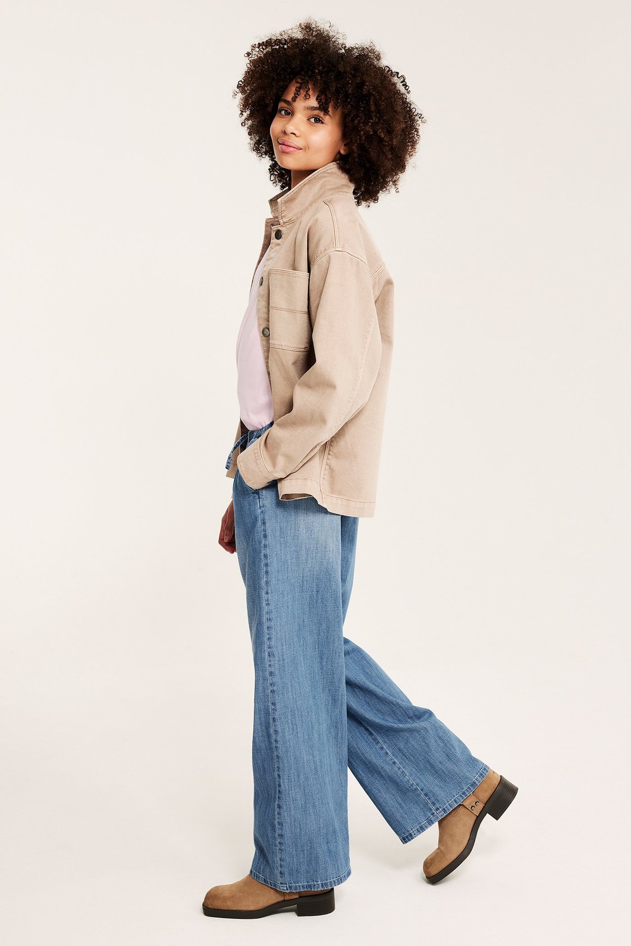 Barn bär ljusblå vida denimbyxor med dragsko, beige jacka, rosa t-shirt och bruna kängor.