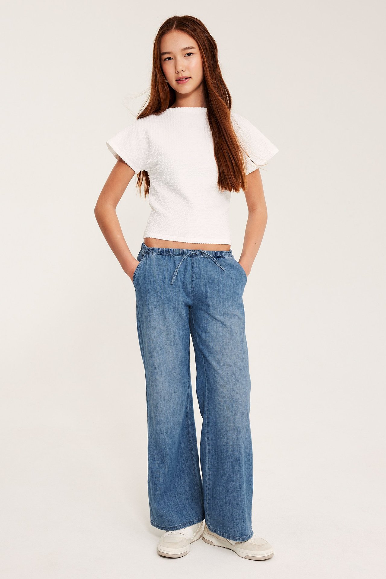 Widok z przodu: Dziecko w jasnych dżinsowych spodniach z szerokimi nogawkami, ze sznurkiem, białym topie i sneakersach.