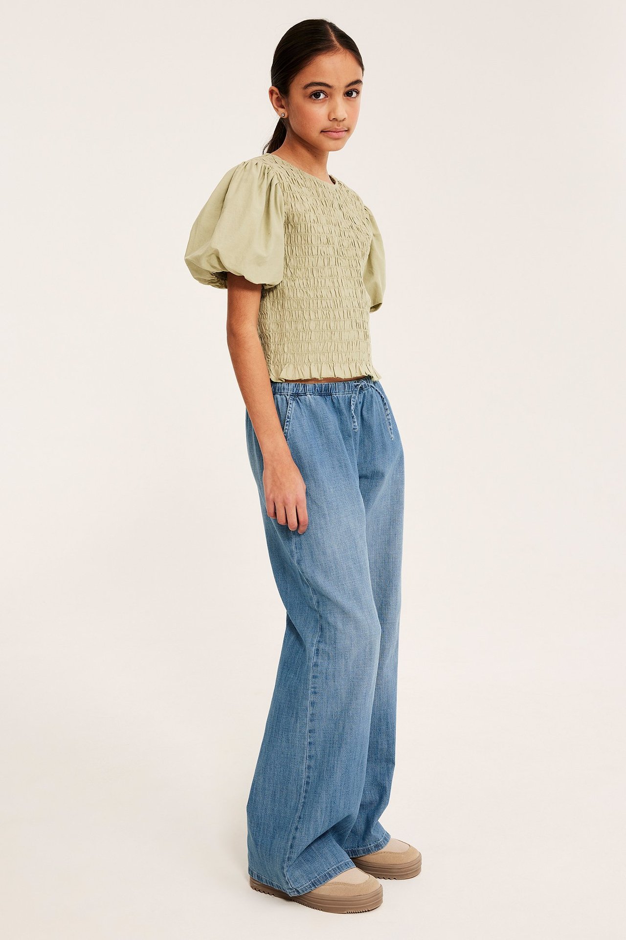 Lapsi käyttää vaaleansinisiä leveälahkeisia nyörifarkkuhousuja, vaaleanvihreää smocktoppia ja beigenvärisiä platform-kenkiä.