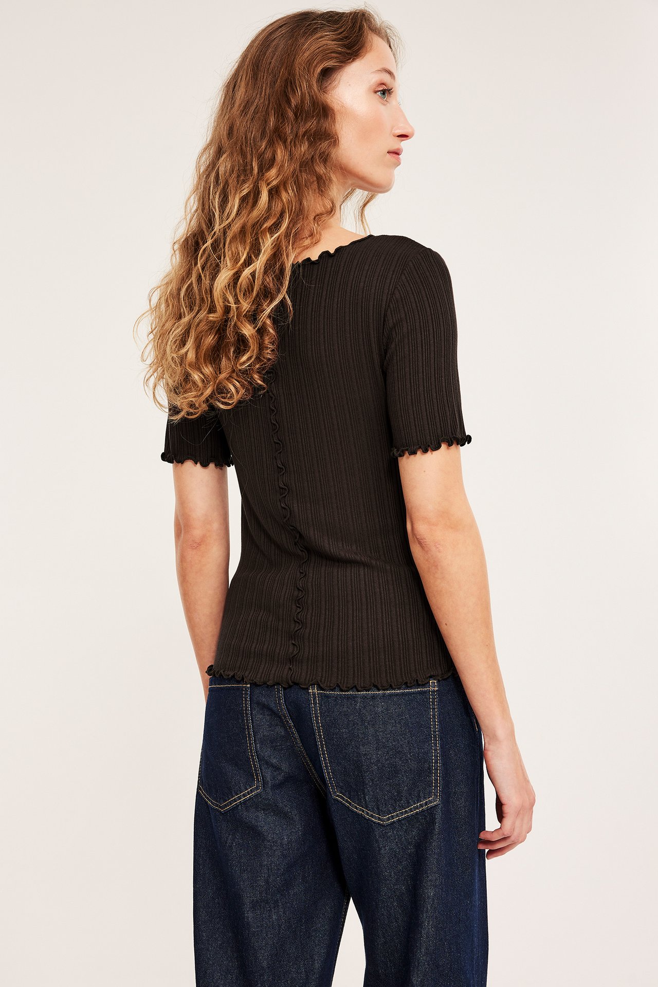 Rygg-sidovy av en kvinna i mörkbrun ribbad kortärmad topp med vågiga sömmar och mörkblå vida jeans.