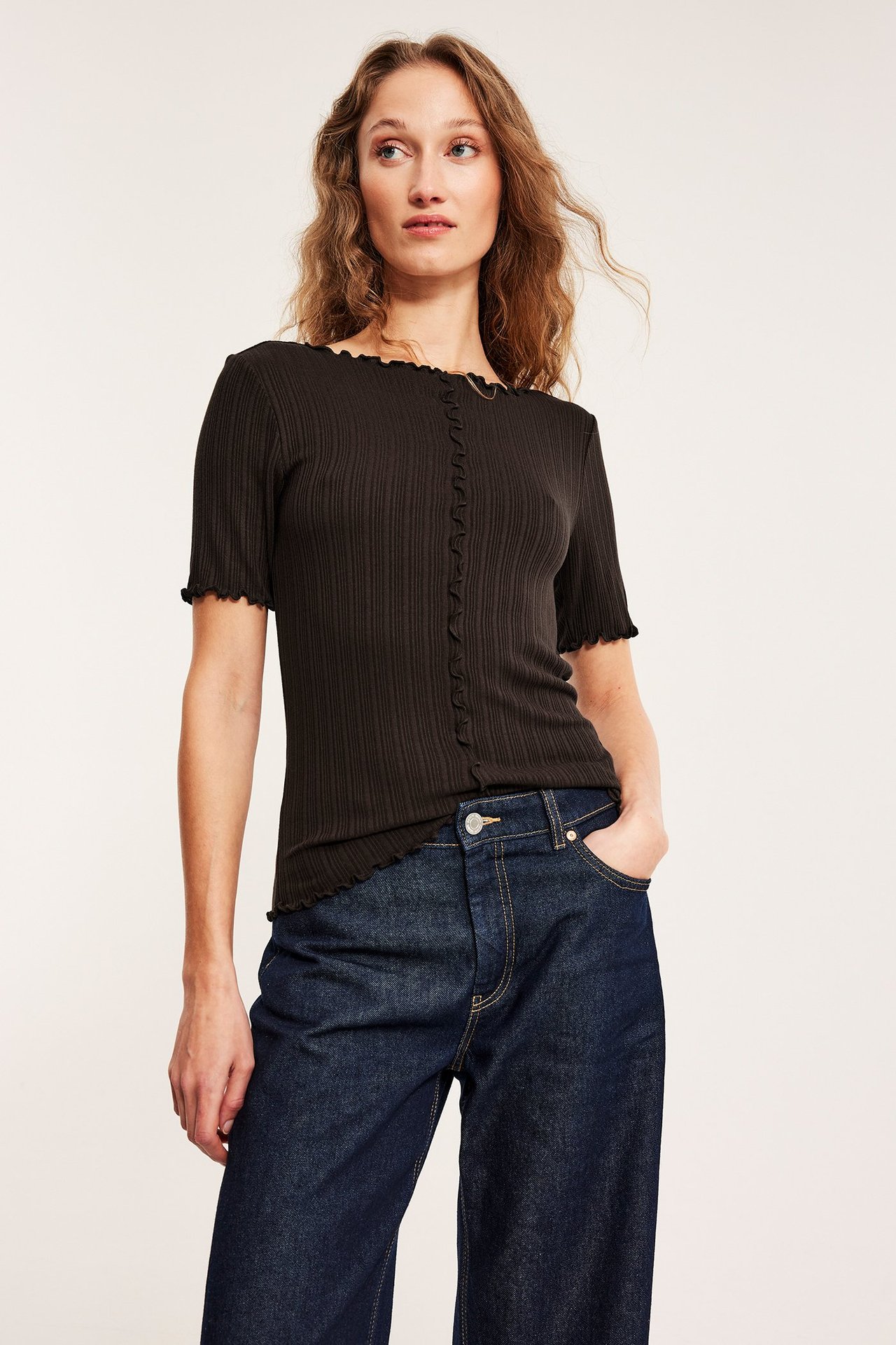 Framifrån, kvinna i mörkbrun ribbad kortärmad topp med babyoverlockdetaljer och mörkblå jeans.