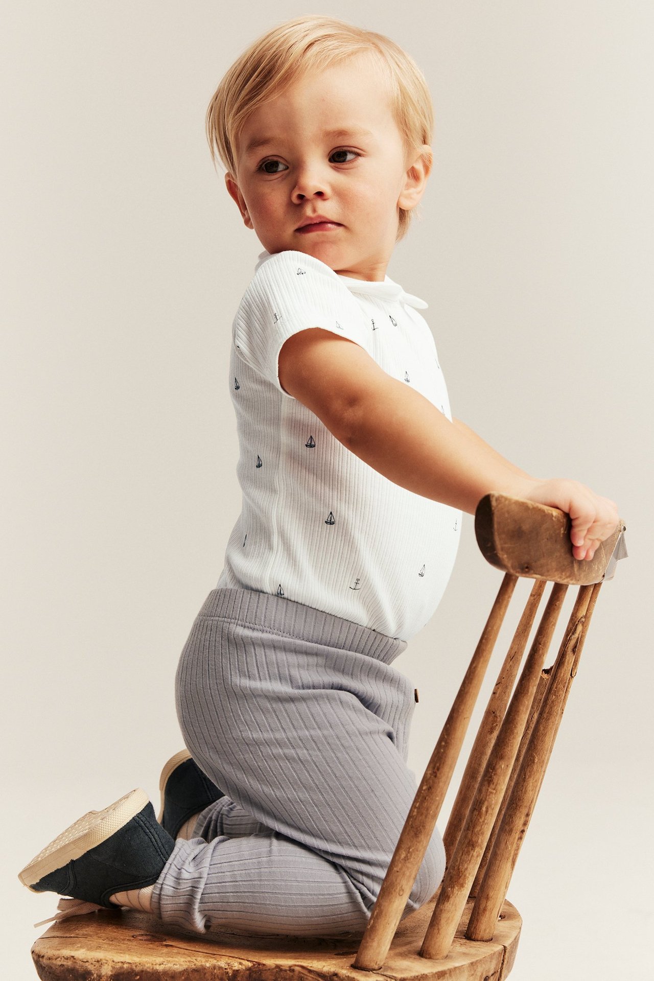 Sidebilde av baby i en offwhite ribbet kortermet body med seilbåtmønster og grå ribbede leggings, knestående.