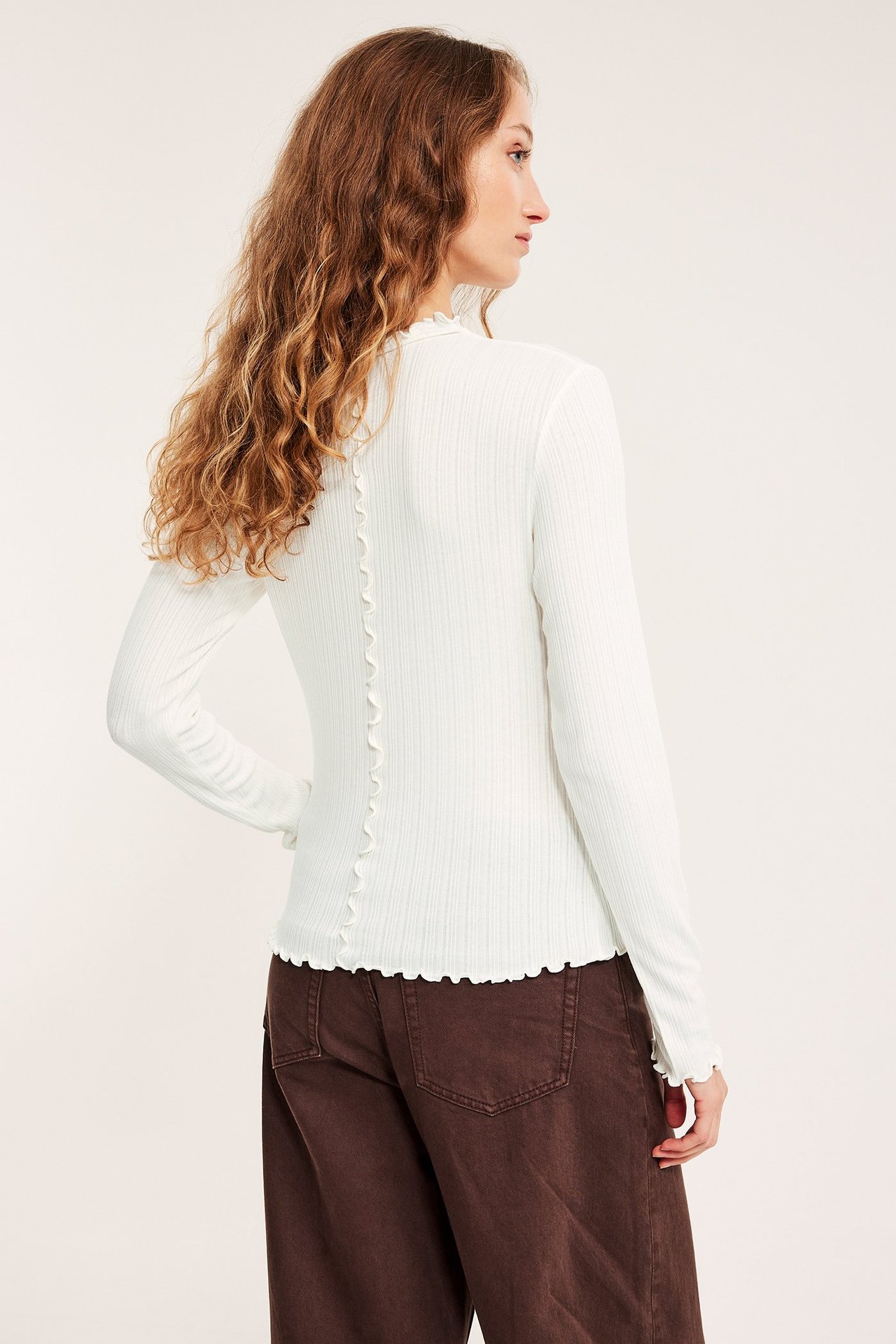 Kvinnans ryggvy i en offwhite ribbad långärmad topp med babyoverlockkanter, stylad med bruna byxor.