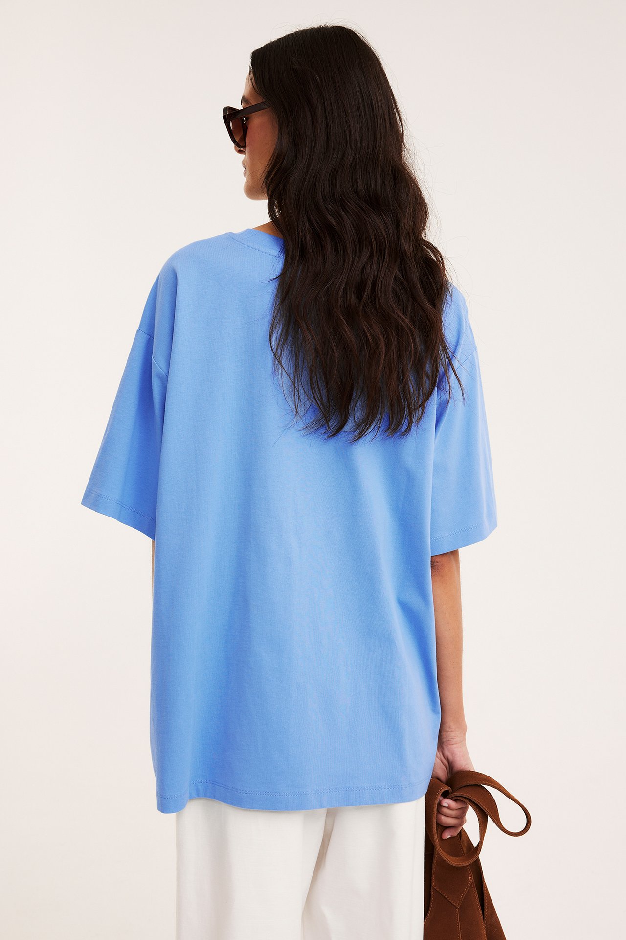 Baksida av kvinna i blå överdimensionerad T-shirt i bomull, vita byxor, mörka solglasögon, håller brun väska.