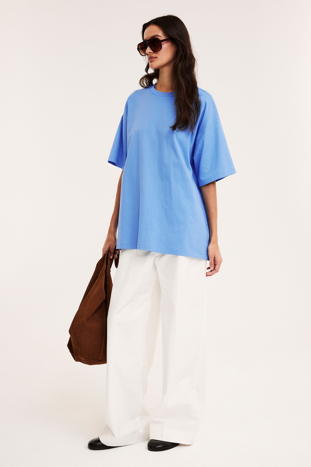 Kvinna, sidovy, i blå oversized t-shirt, vita byxor, svarta skor, håller brun tygväska, bär solglasögon.