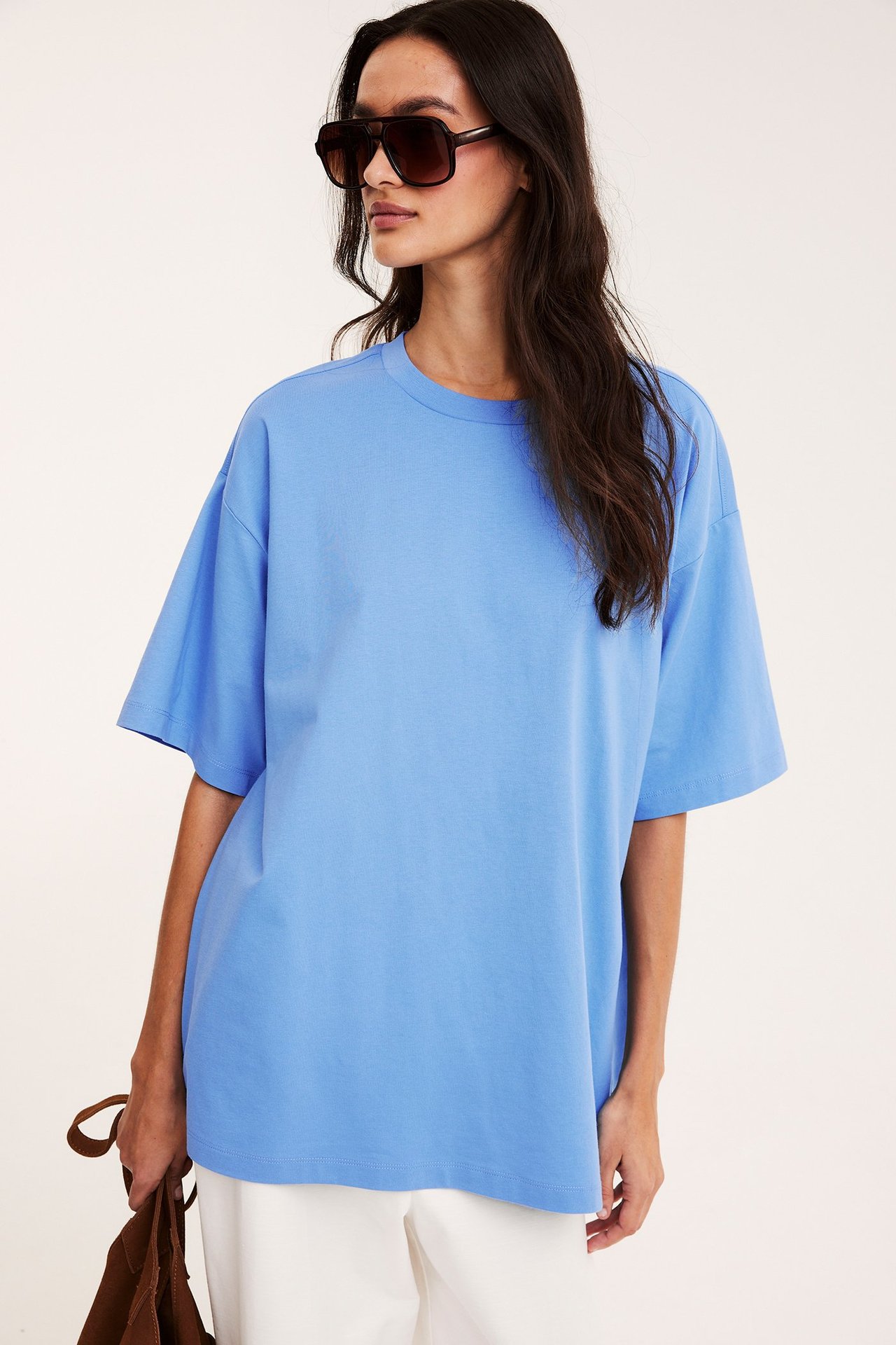 Kobieta w jasnoniebieskim T-shircie oversize, białych spodniach, ciemnych okularach, z brązową torbą.
