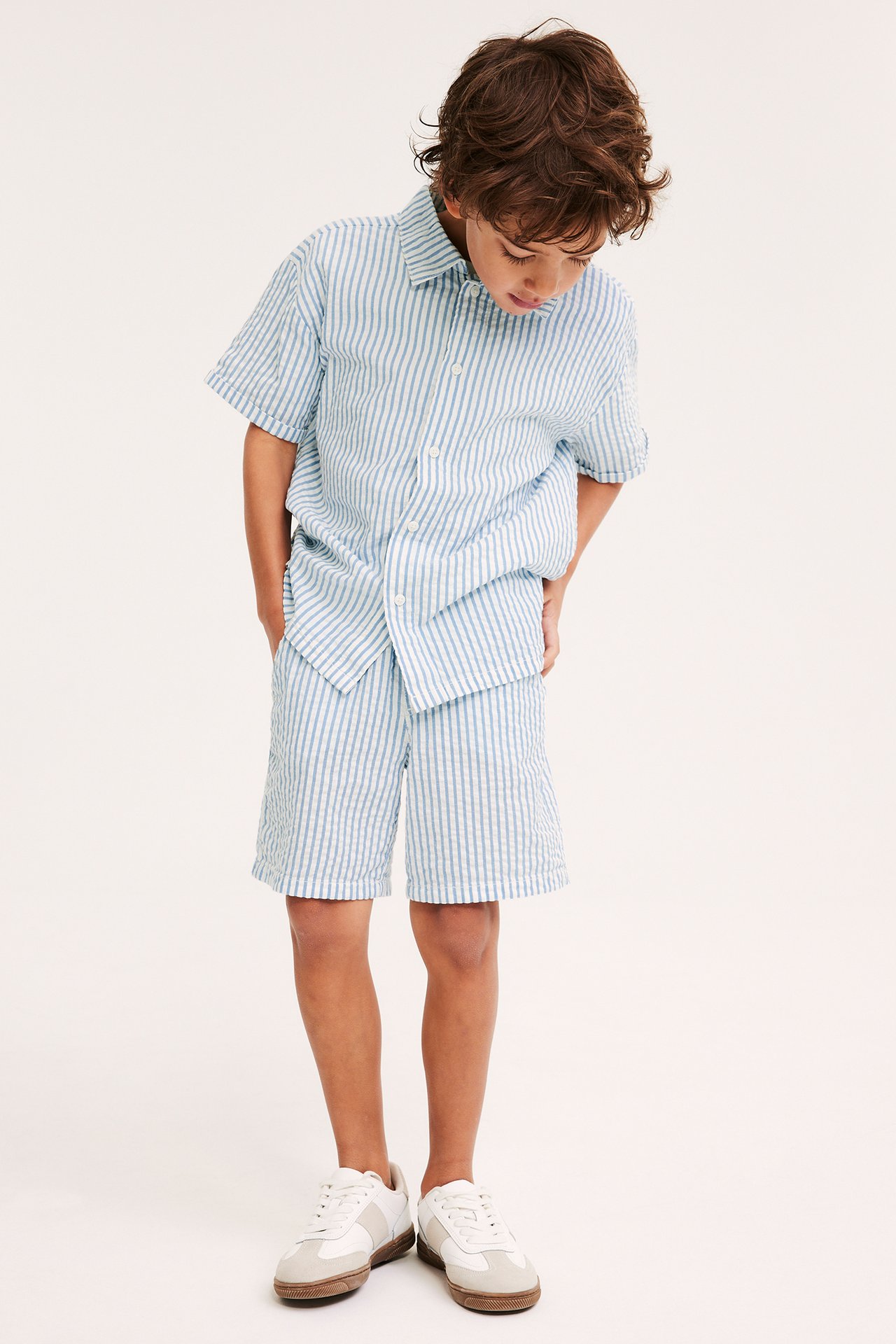 Etunäkymä lapsesta vaaleansinisessä raidallisessa paitassa, yhteensopivissa shortseissa ja tennareissa.