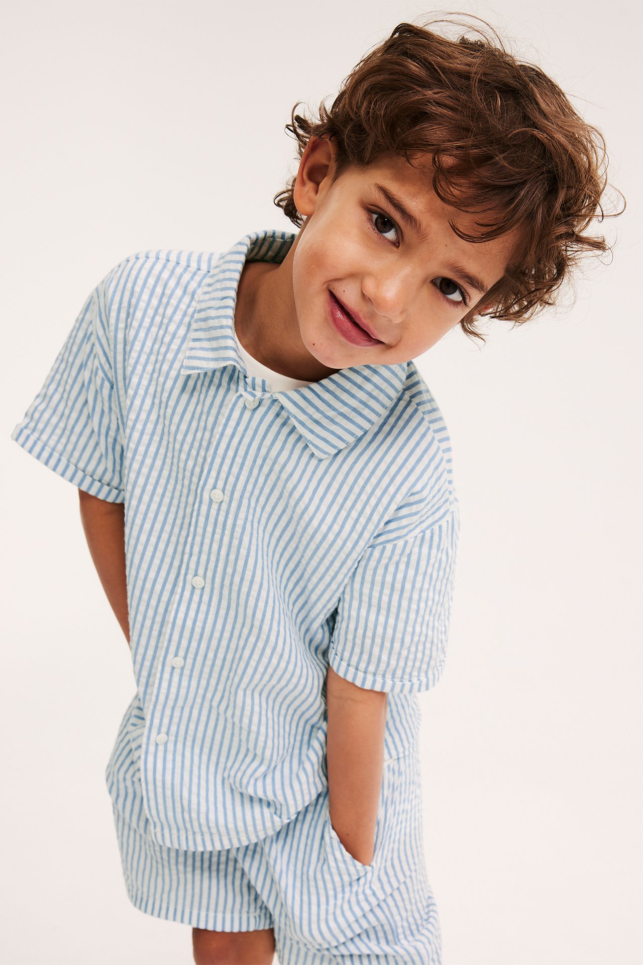 Framifrån, ett barn i blå- och vitrandig kortärmad bomullsseersucker-skjorta och matchande shorts.