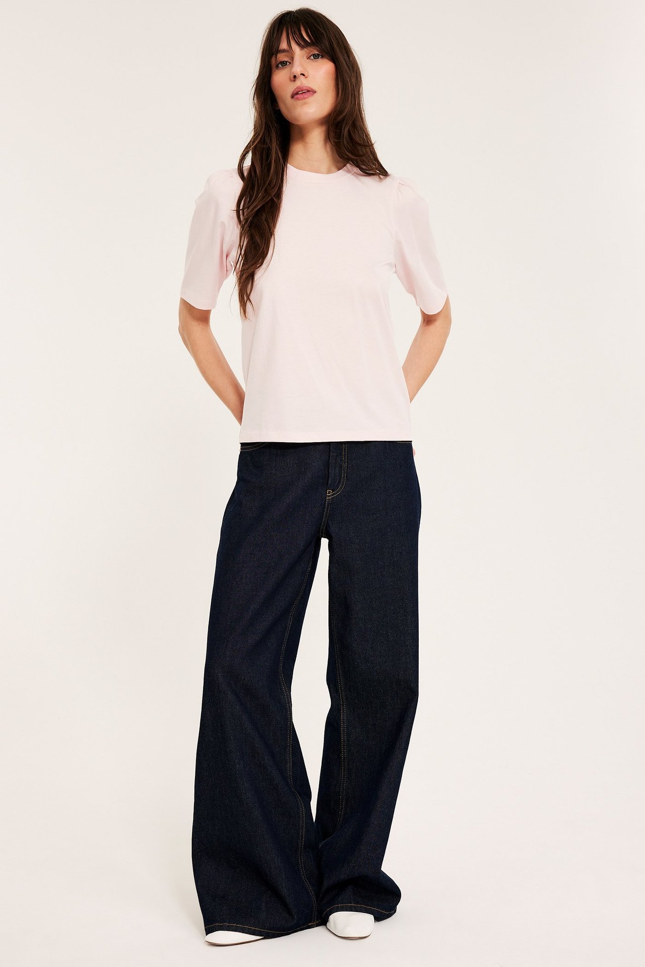 Forfra, kvinne i lys rosa bomullstopp med puffermer, mørke vide jeans og hvite sko.