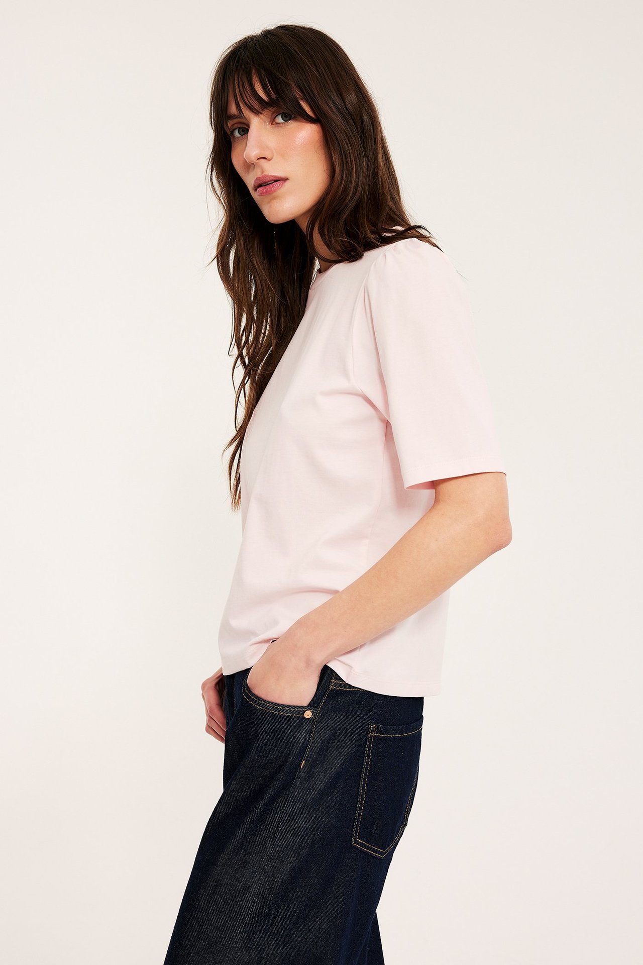 Sidebilde av kvinne i lyserosa bomullstopp med puffermer og mørkeblå jeans.