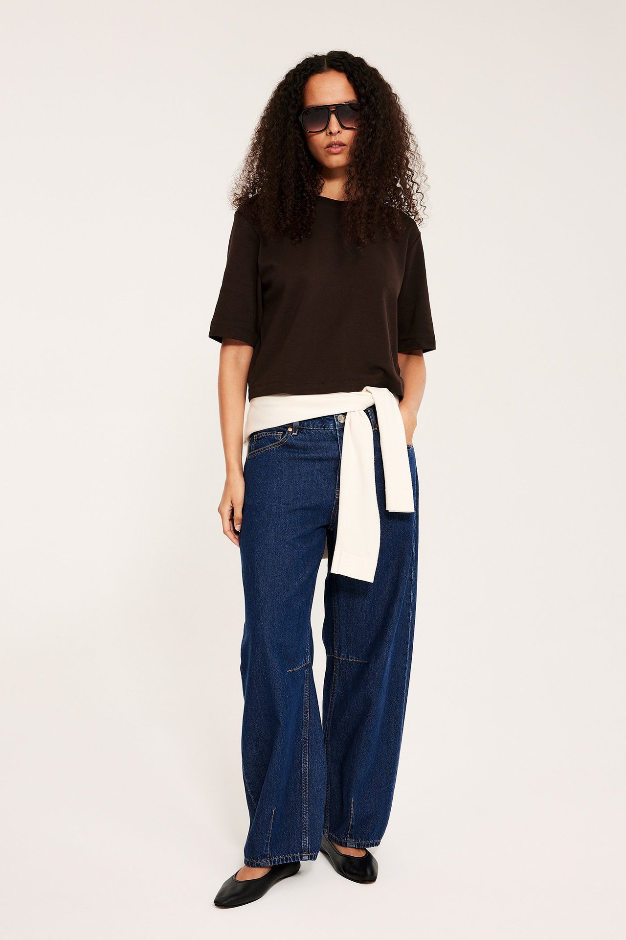 Edestä: Nainen tummanruskea lyhyt t-paita, siniset farkut, valkoinen neule, mustat kengät.