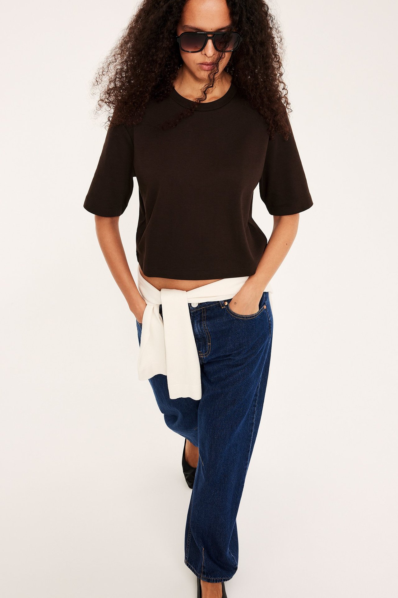 Nainen yllään tummanruskea lyhyt t-paita, tummansiniset farkut, aurinkolasit ja valkoinen pusero vyötäröllä.