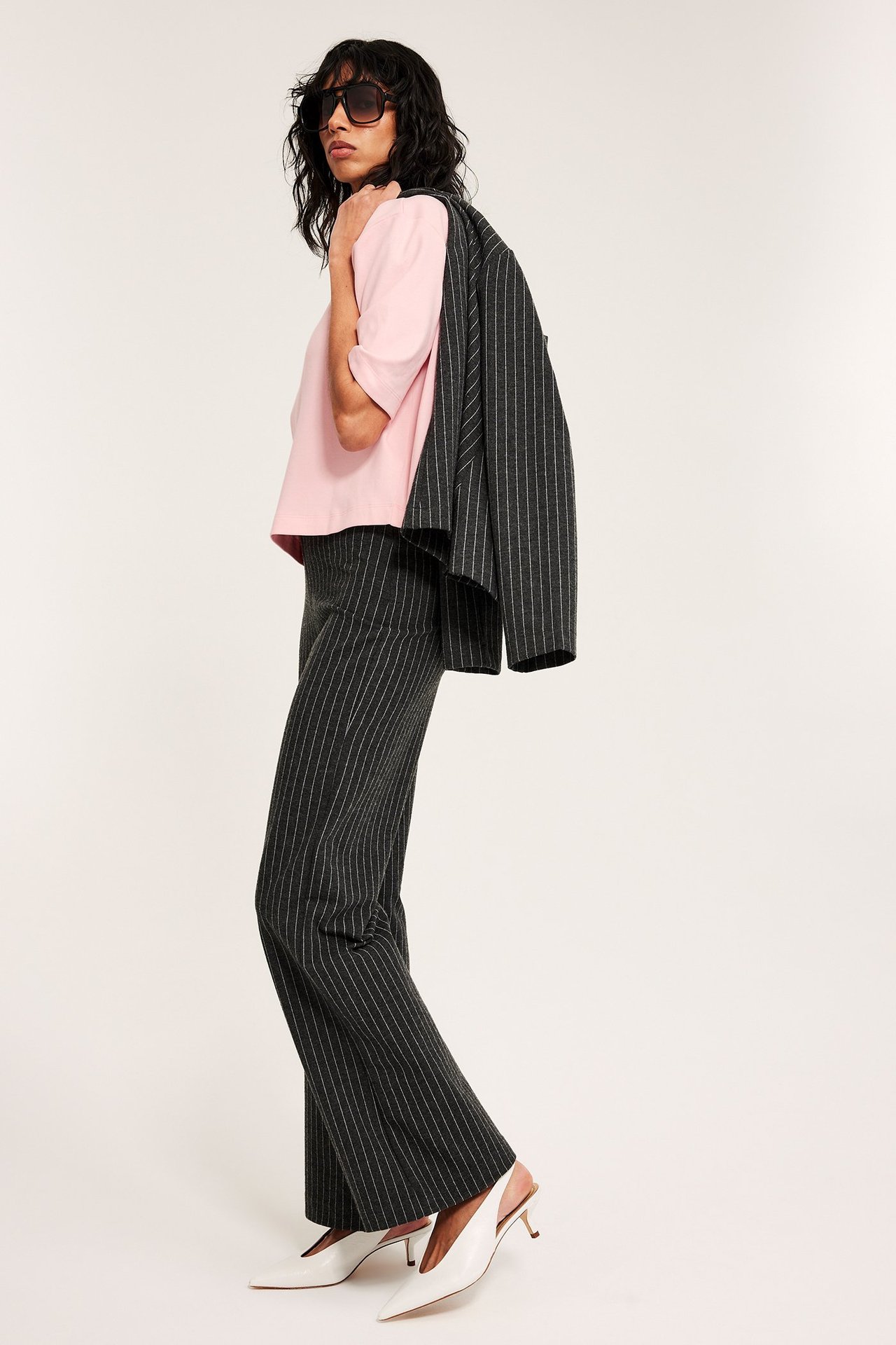 Nainen sivulta: vaaleanpunainen cropattu puuvillainen t-paita, tummat raidalliset housut, bleiseri olalla, valkoiset korot.