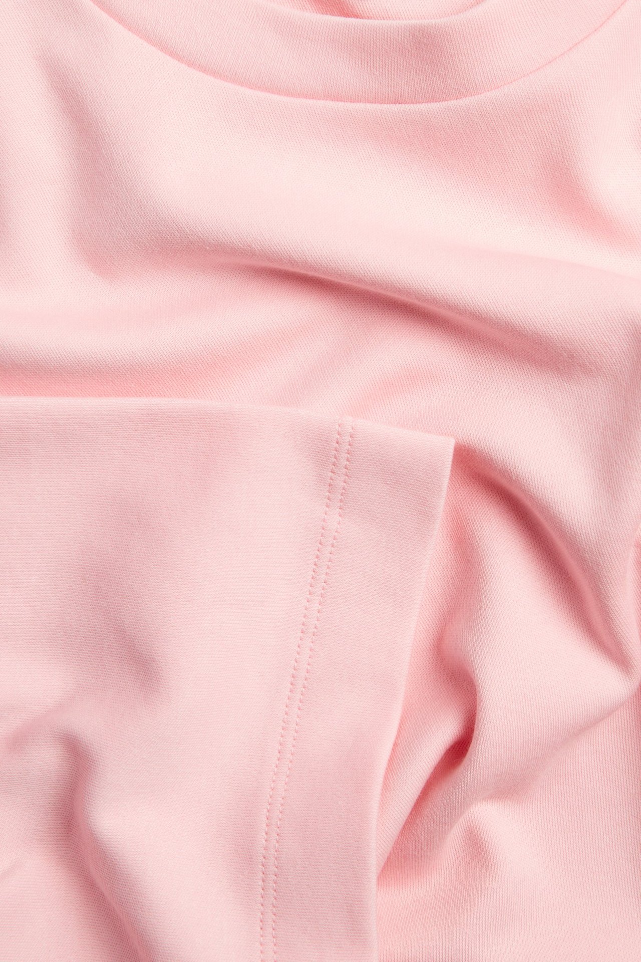 Zbliżenie jasnoróżowego bawełnianego materiału koszulki, widoczna faktura, dekolt i przeszyty szew.