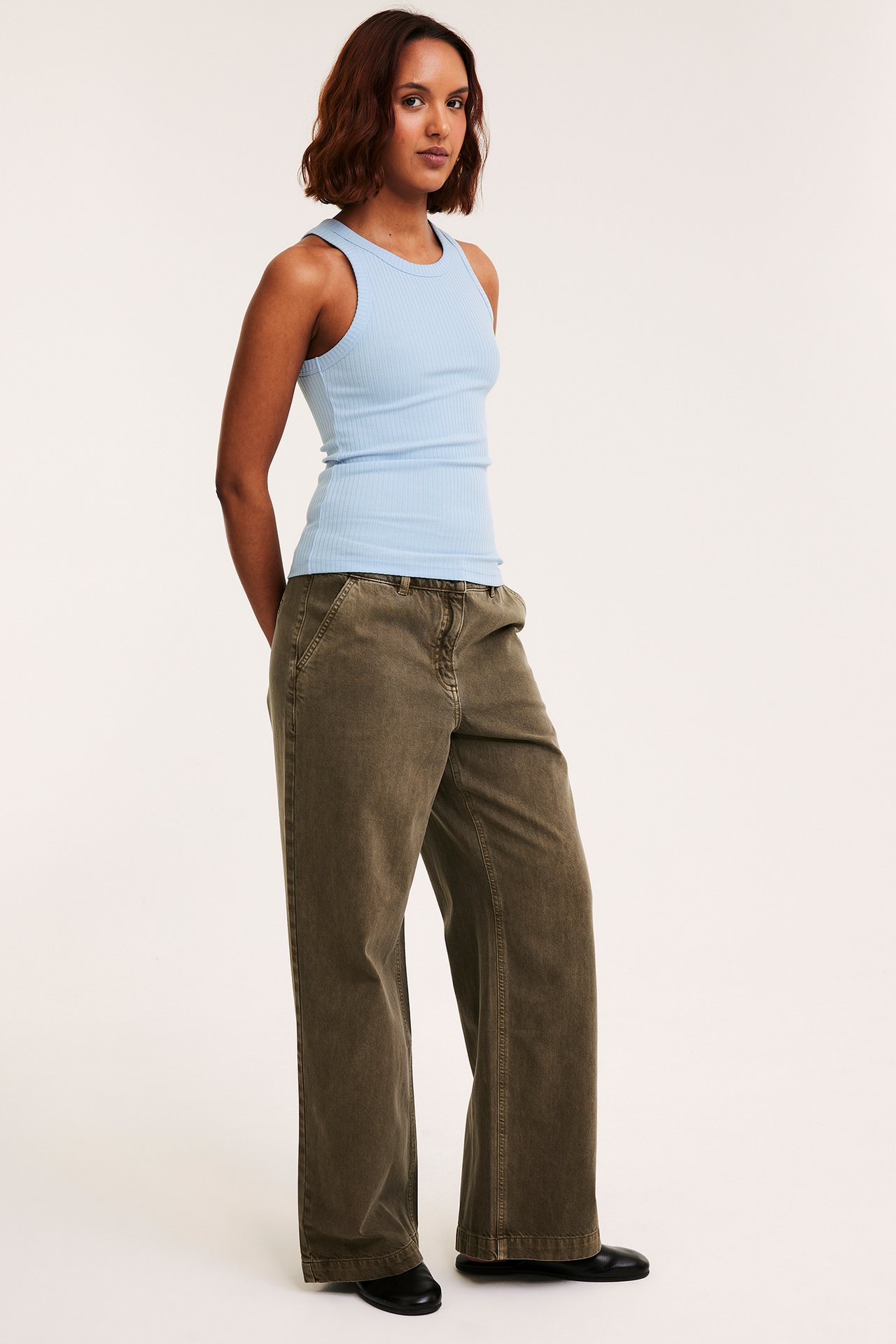 Sidovy av kvinna i ljusblått ribbat linne, mörkgröna vida byxor och svarta platta skor.