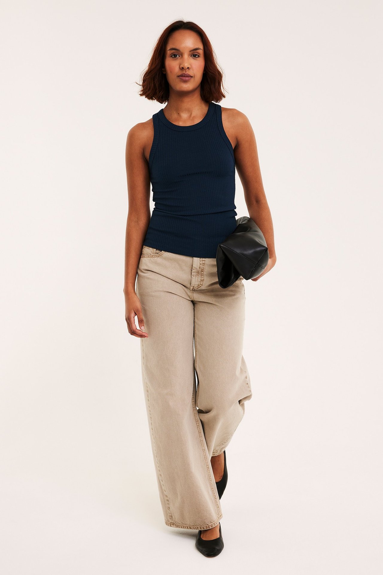 Frontvy: kvinna i mörkblått ribbat linne, beiga vida byxor, svarta skor, med svart clutch.