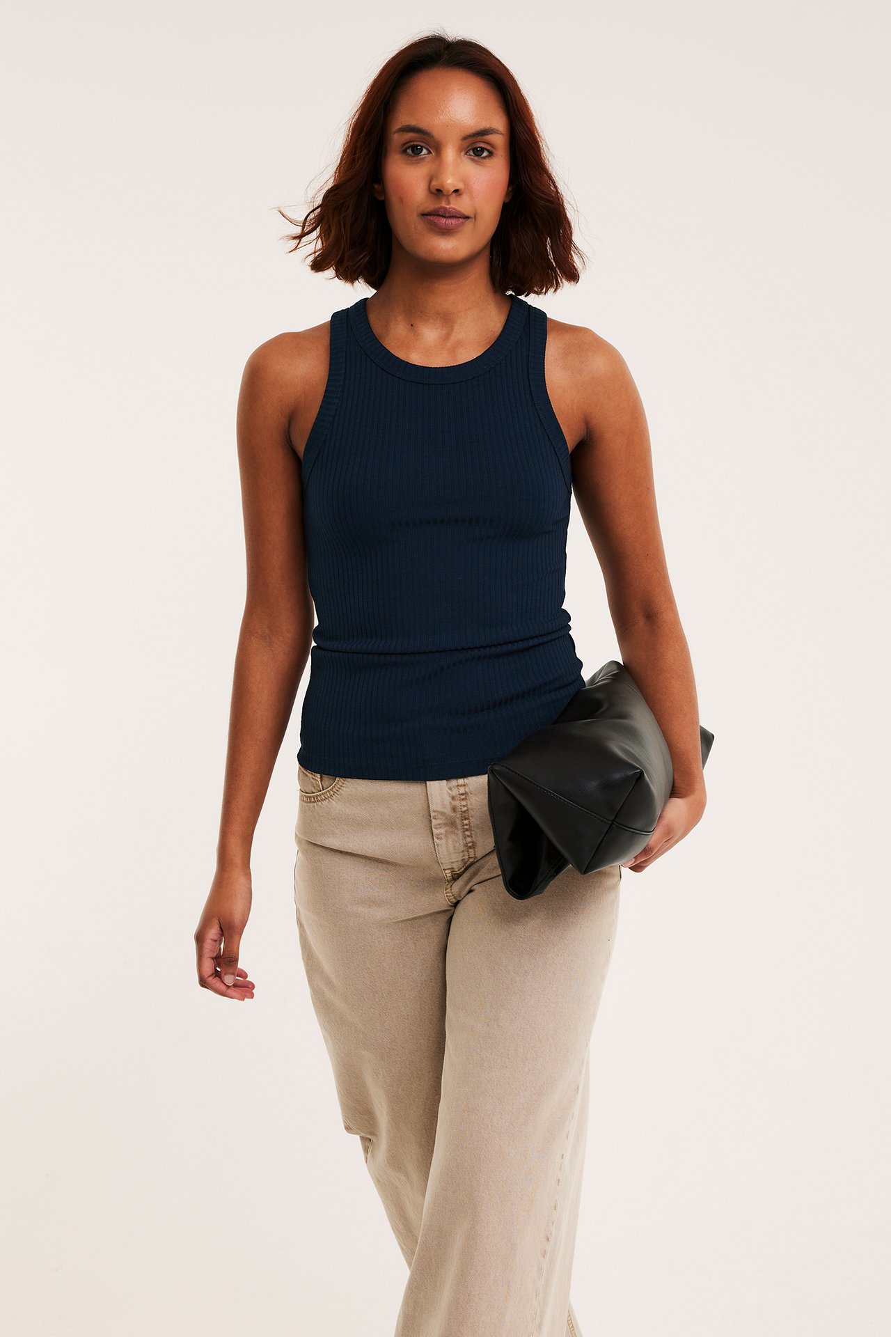 Framifrån, en kvinna i mörkblått ribbat linne, ljusbeige vida byxor, håller en svart clutchväska.