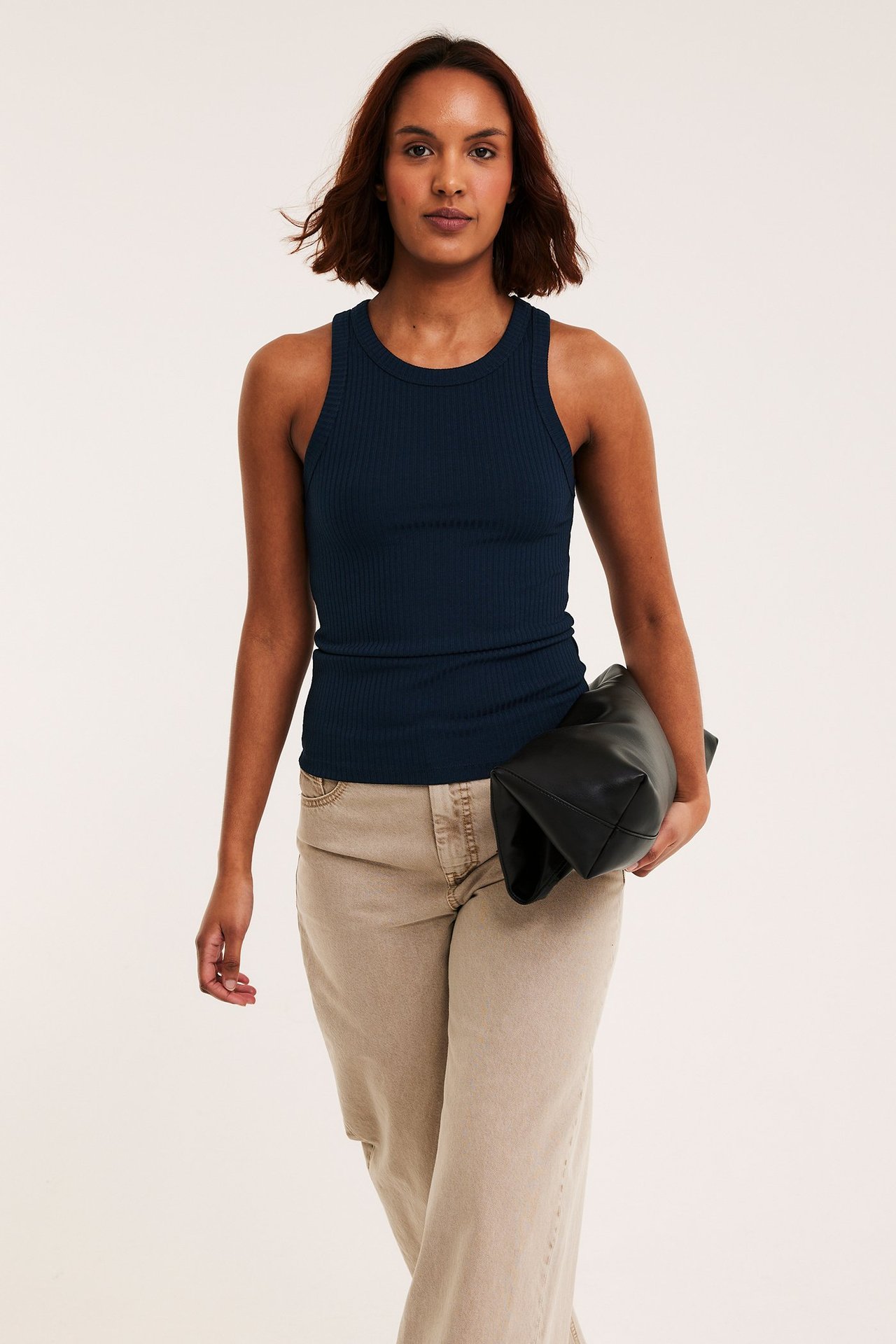 Edestä, nainen tummansinisessä ribbitoppissa, vaaleanbeigeissä leveälahkeisissa housuissa, pitäen mustaa clutch-laukkua.