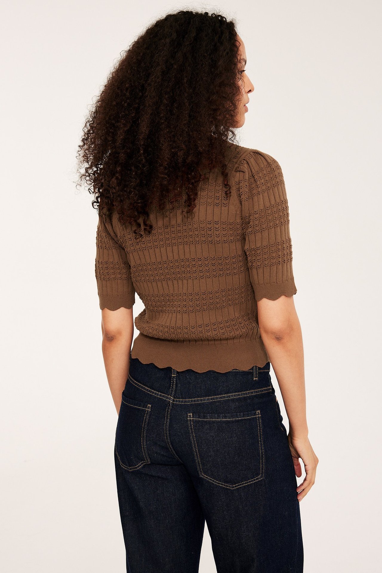 Ryggvy av kvinna i brun hålstickad kortärmad bomullstopp med vågiga kanter och mörkblå jeans.