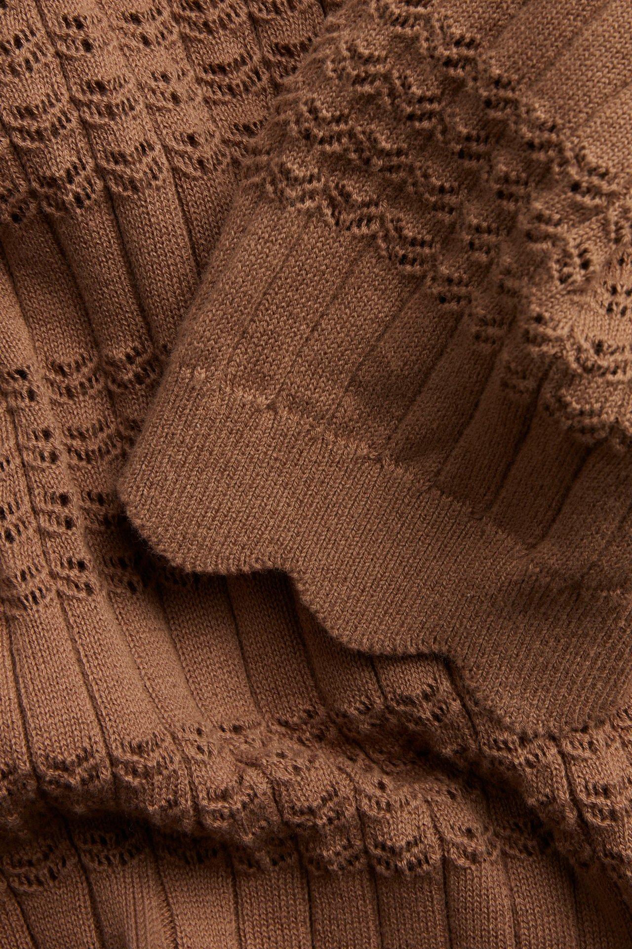 Närbild av en brun kortärmad stickad topp i bomull med ribbade partier, hålmönster och vågig kant.